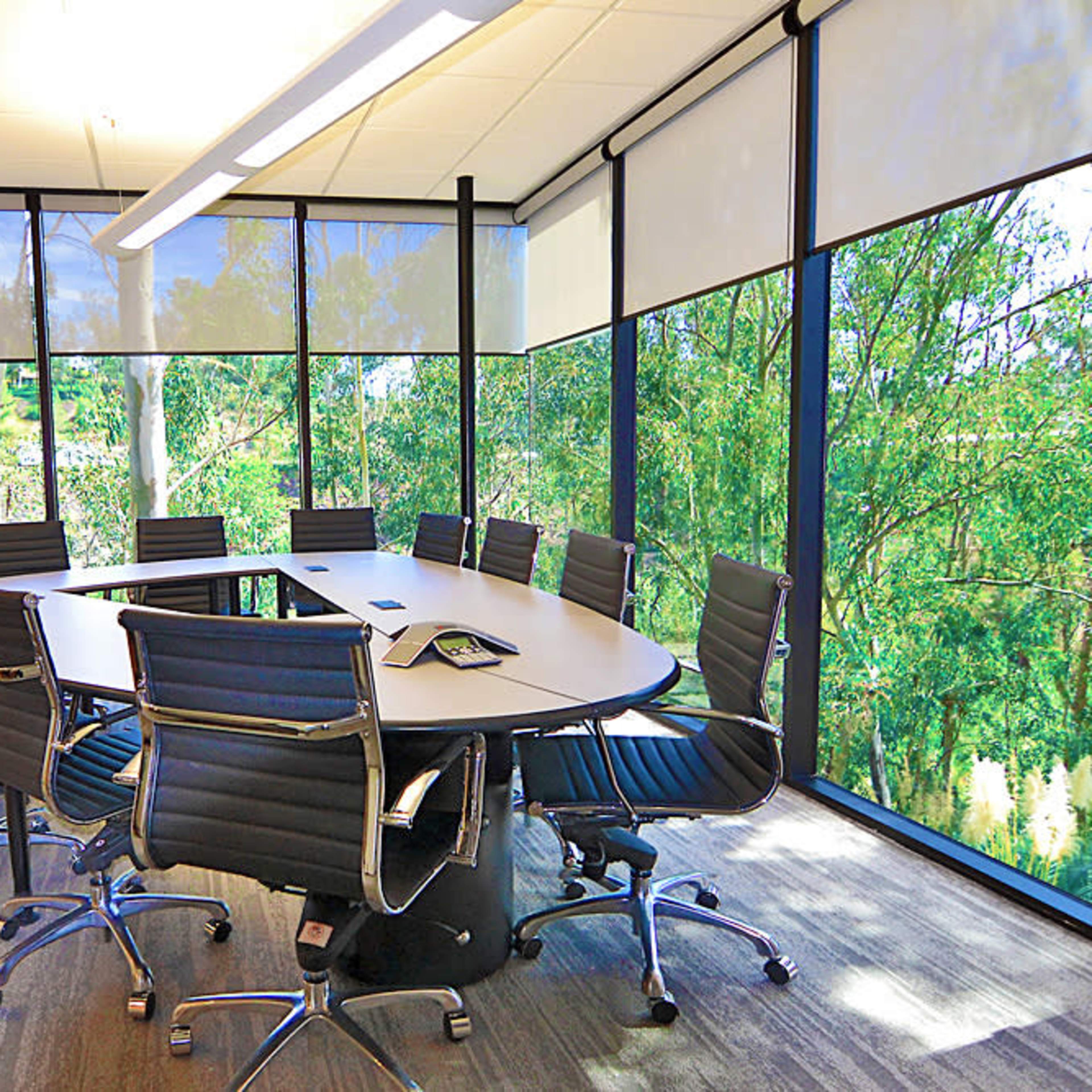 A conference room features a large oval table surrounded by black chairs and floor-to-ceiling windows showcasing greenery outside.