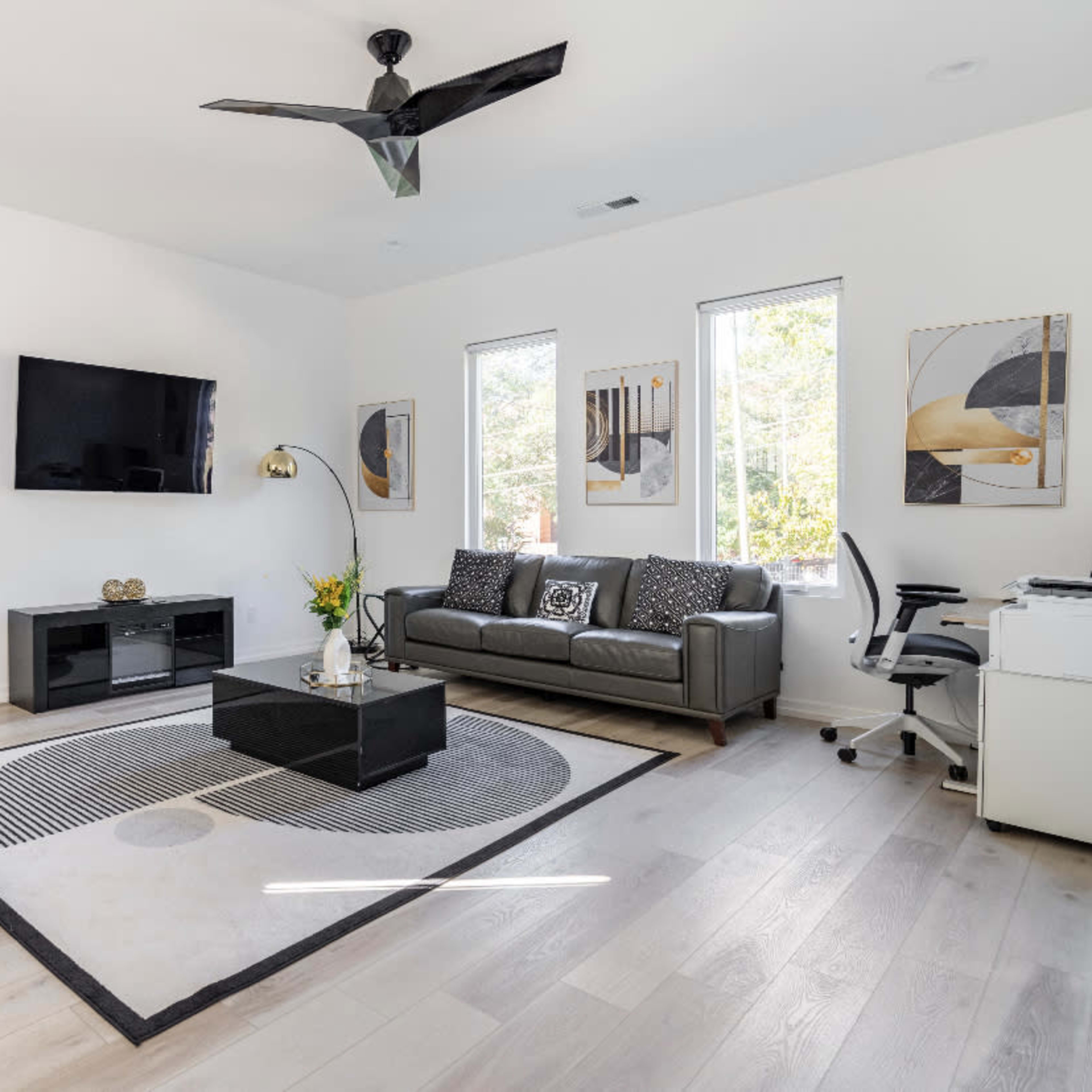 The living room features a large area rug, a black coffee table, a wall-mounted television, and modern furniture arranged around a window with natural light.