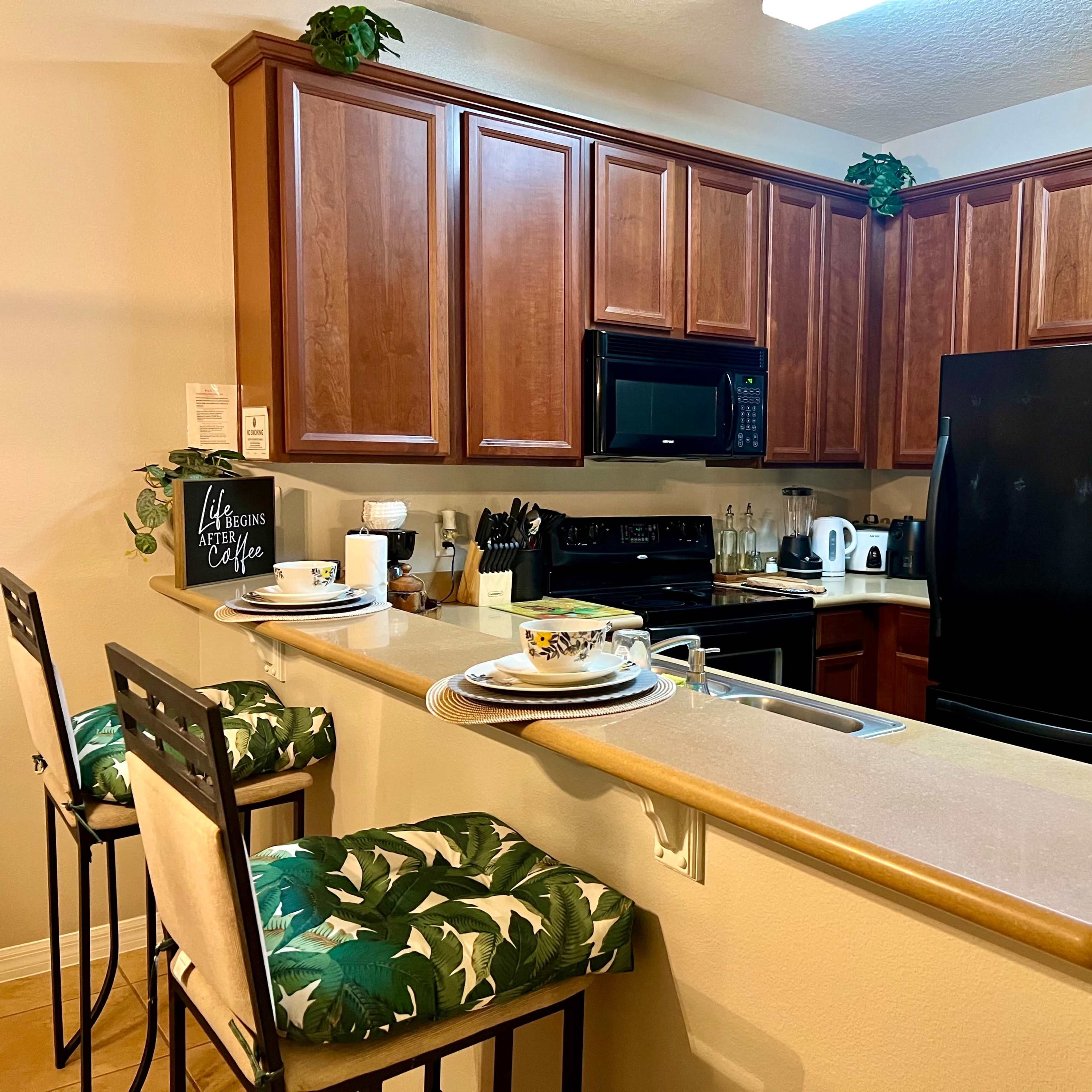 A modern kitchen with wooden cabinetry, black appliances, and bar stools with green tropical patterns.