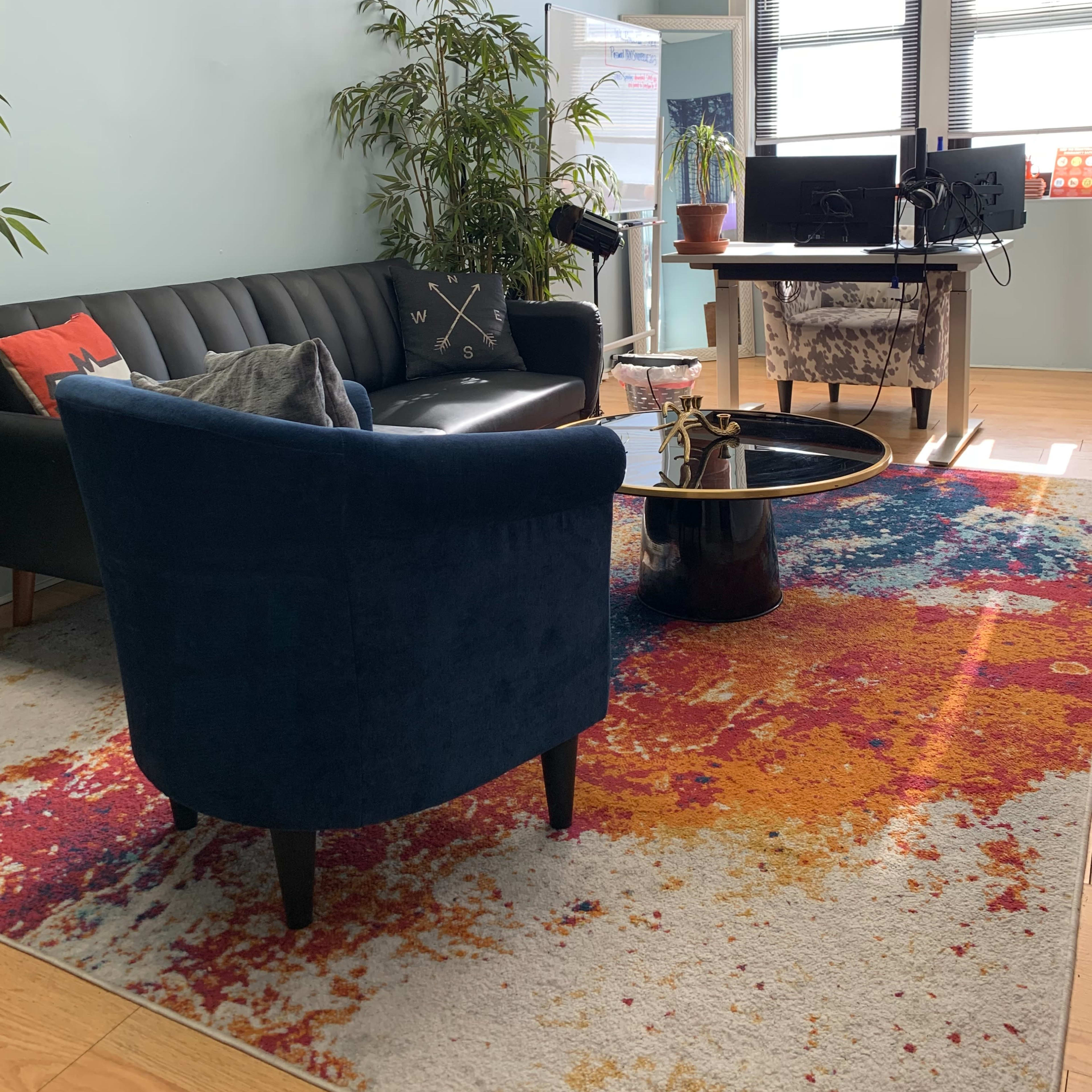 A modern office space with a blue couch, a chair, a round coffee table, and a desk with two monitors, all arranged on a colorful rug.