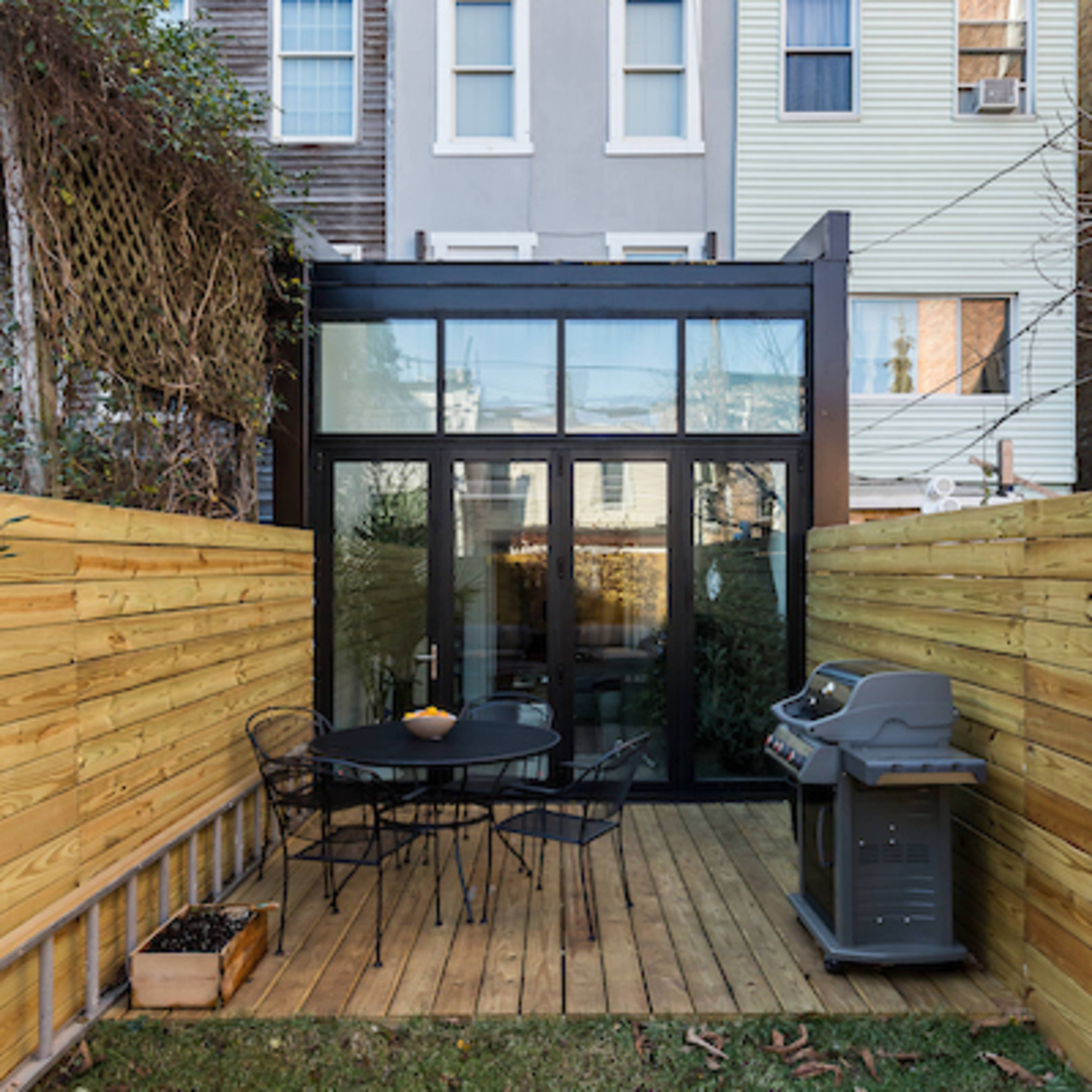 A small outdoor patio with wooden decking, black metal furniture, and a grill, surrounded by wooden fences and adjacent to multi-colored townhouses.