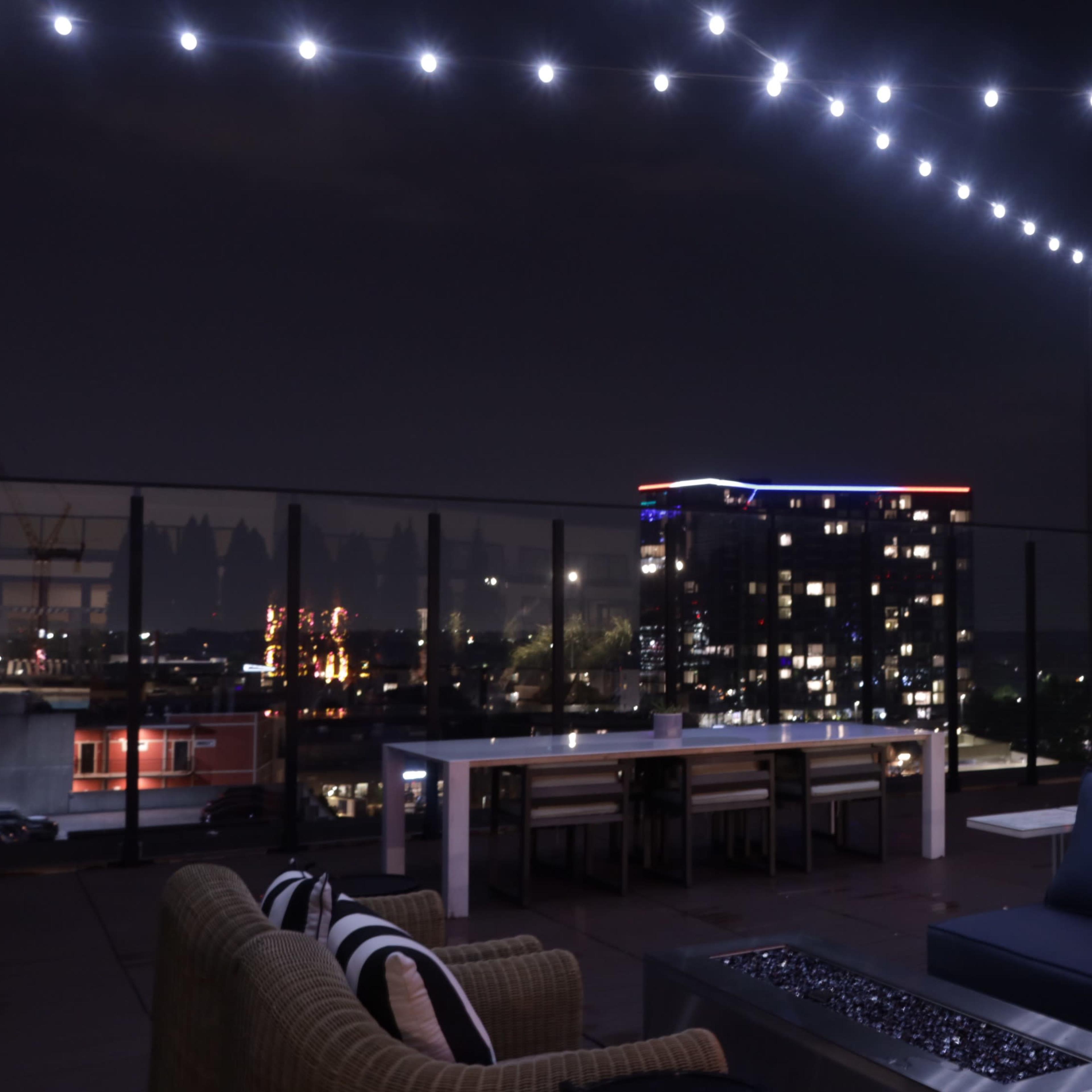 A rooftop terrace at night features string lights overhead, seating arrangements, and a view of illuminated buildings in the distance.