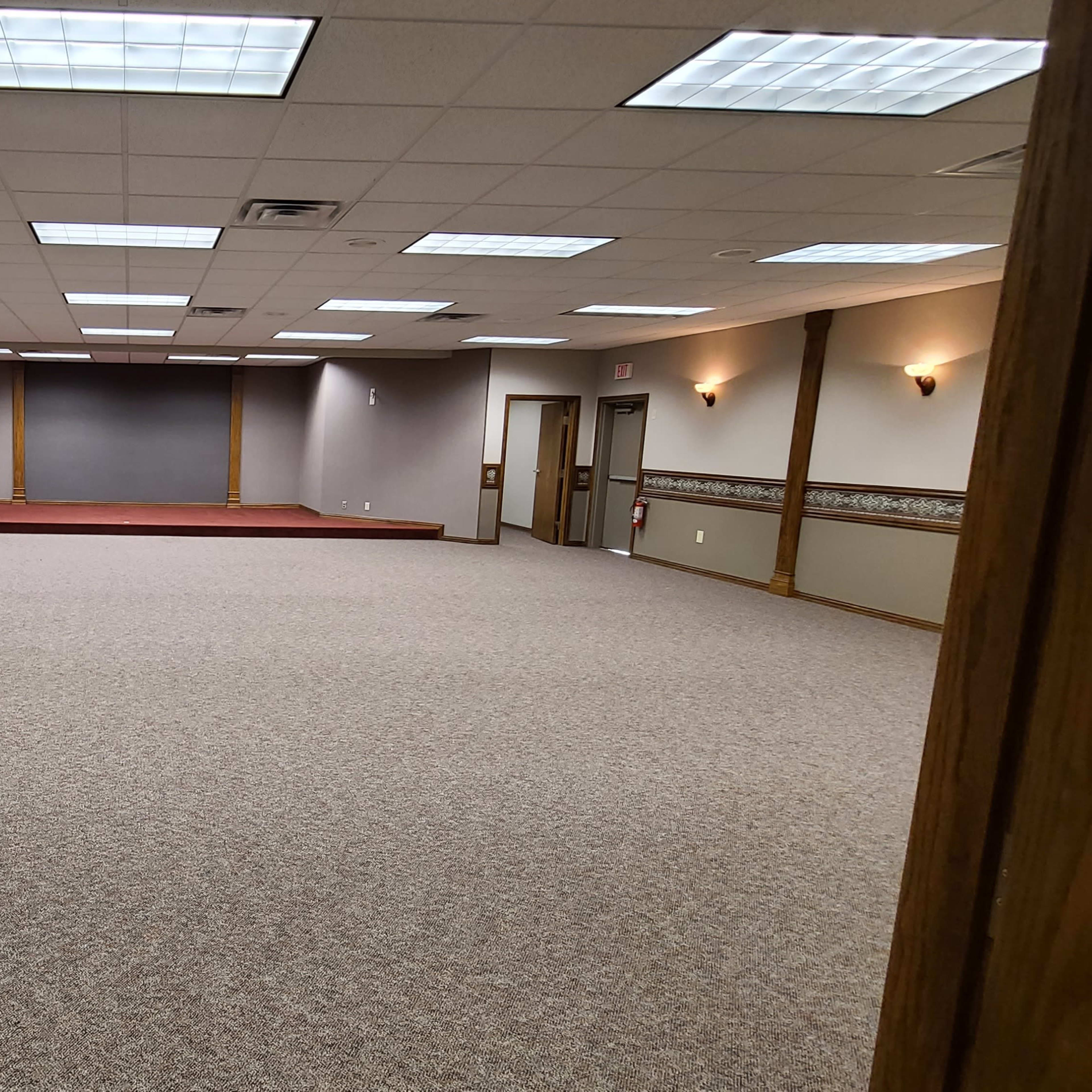 A large, empty room with carpet flooring and a raised stage at the back, illuminated by overhead lights.