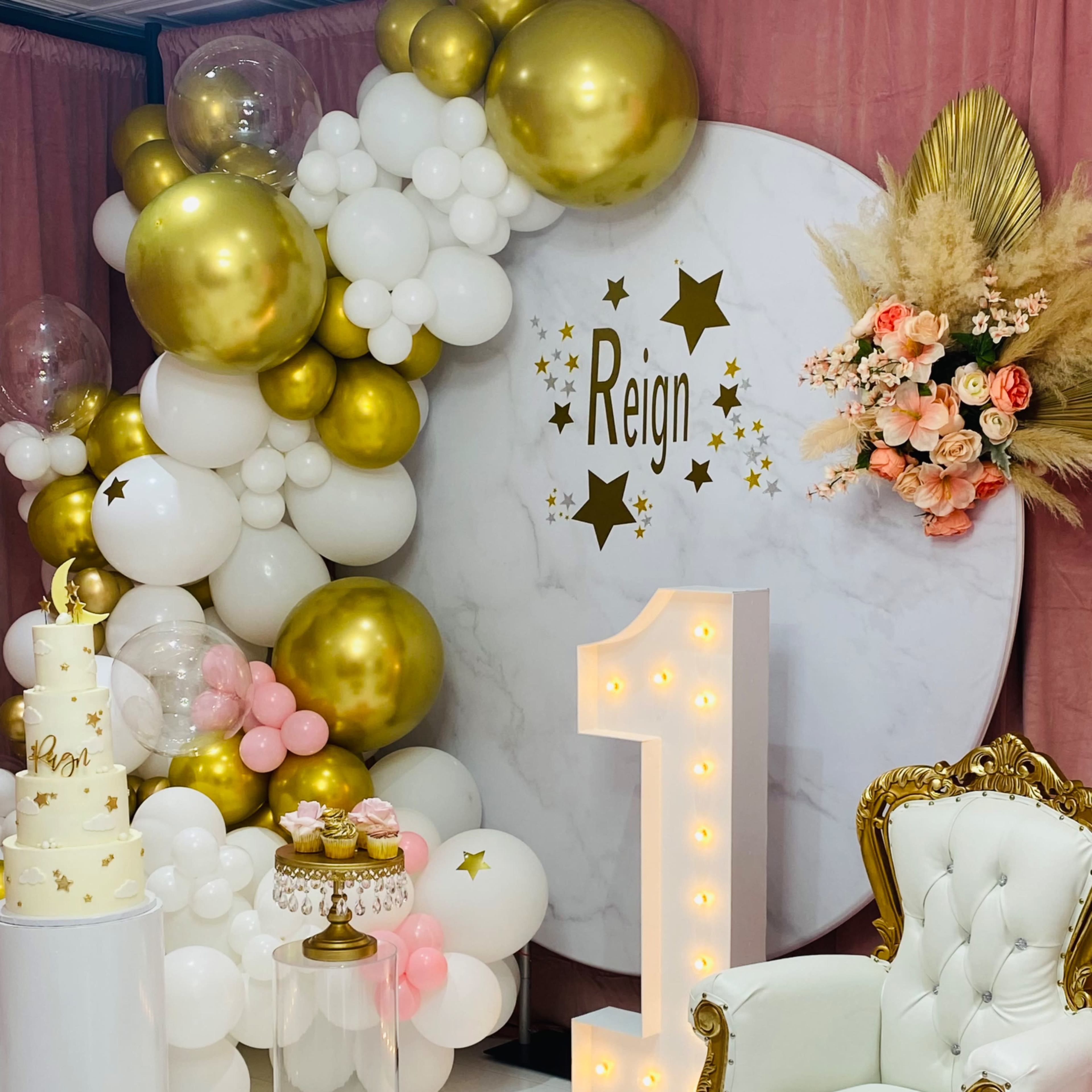 The scene features a decorated area for a first birthday celebration, highlighted by a large circular backdrop, a white and gold cake, and a decorative golden chair surrounded by balloon arrangements and floral accents.