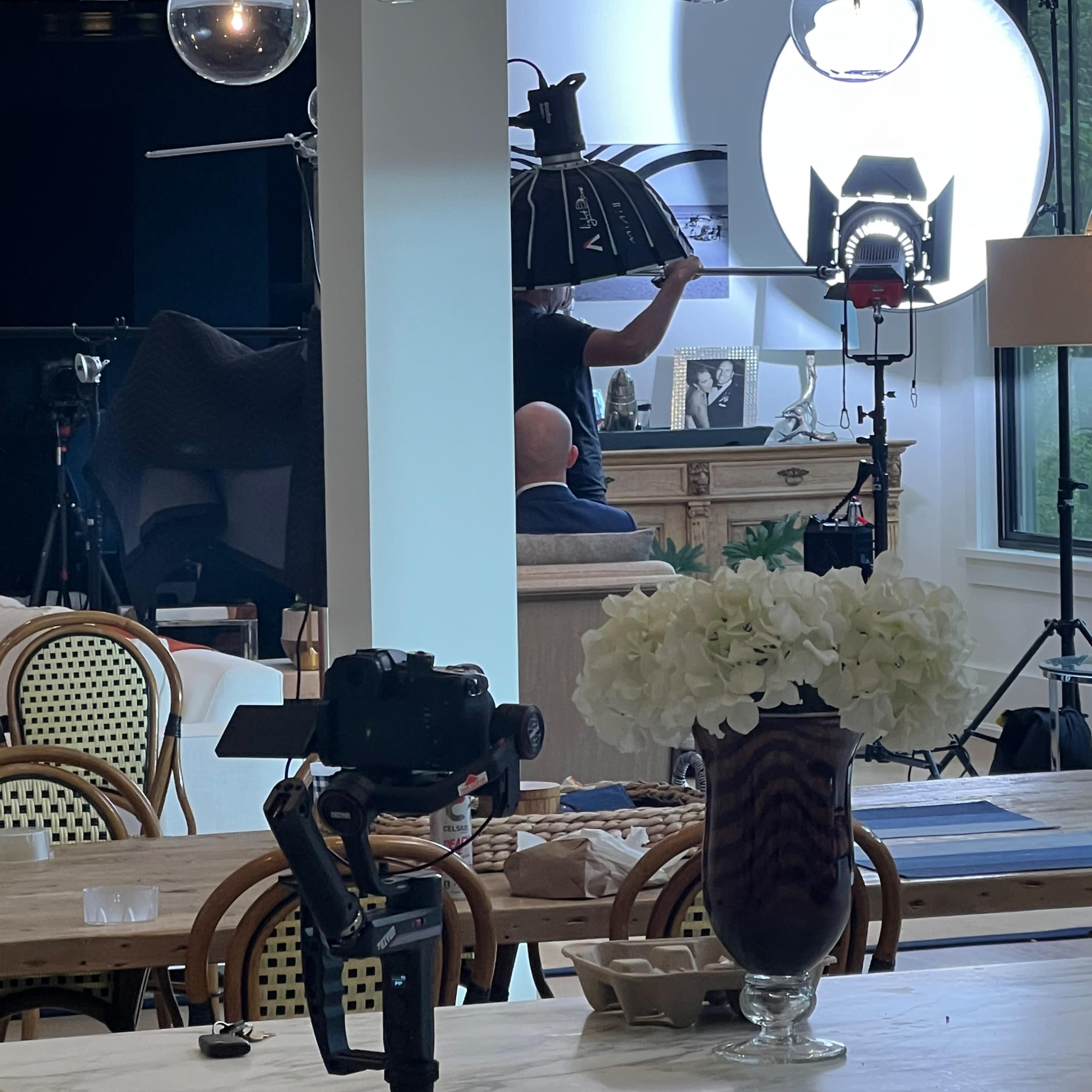 A photography setup is visible in a well-lit room, featuring a photographer with lights and equipment directed toward a subject near a decorative piece.