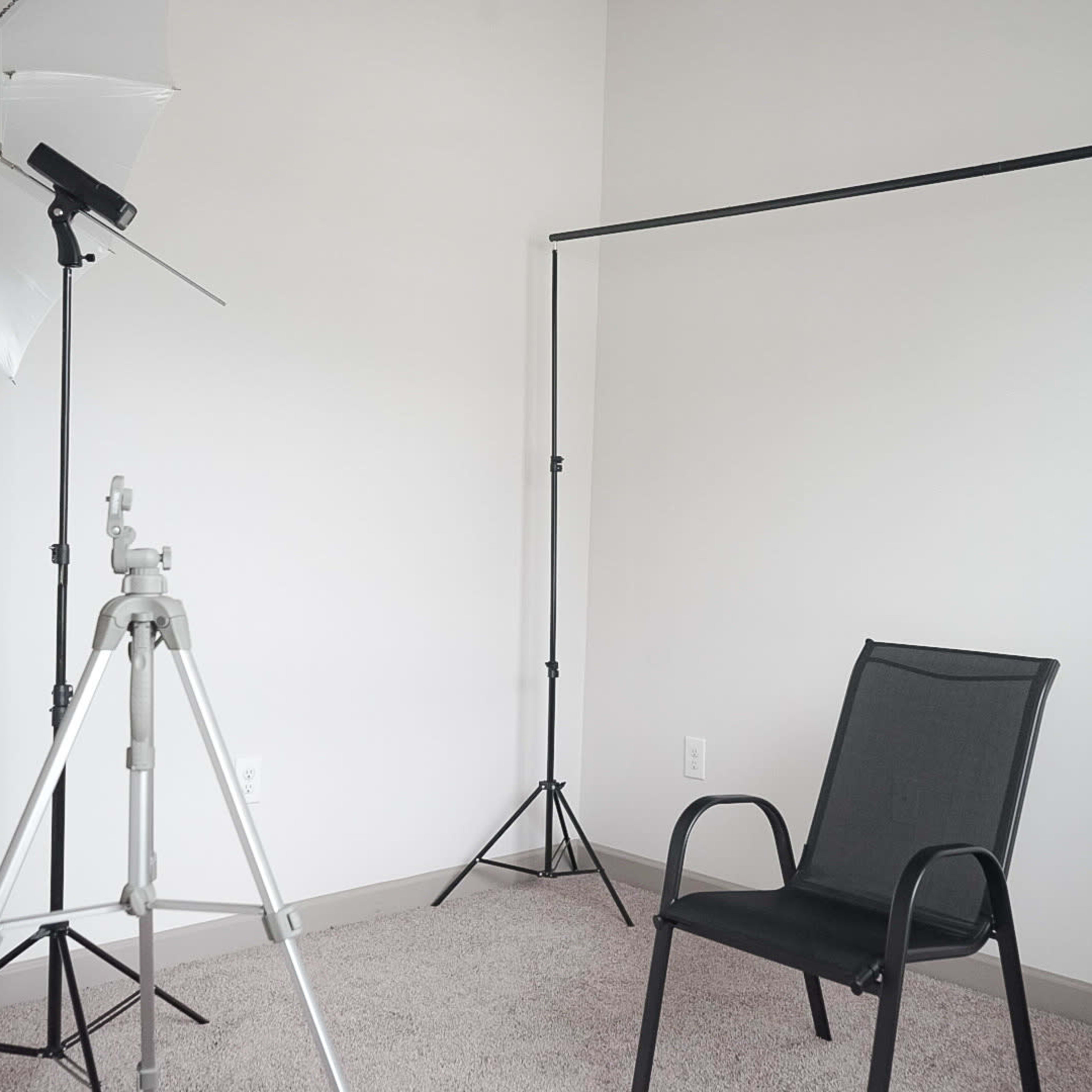 A minimalistic studio setup with a black chair positioned on carpet, accompanied by a lighting umbrella and a backdrop stand.