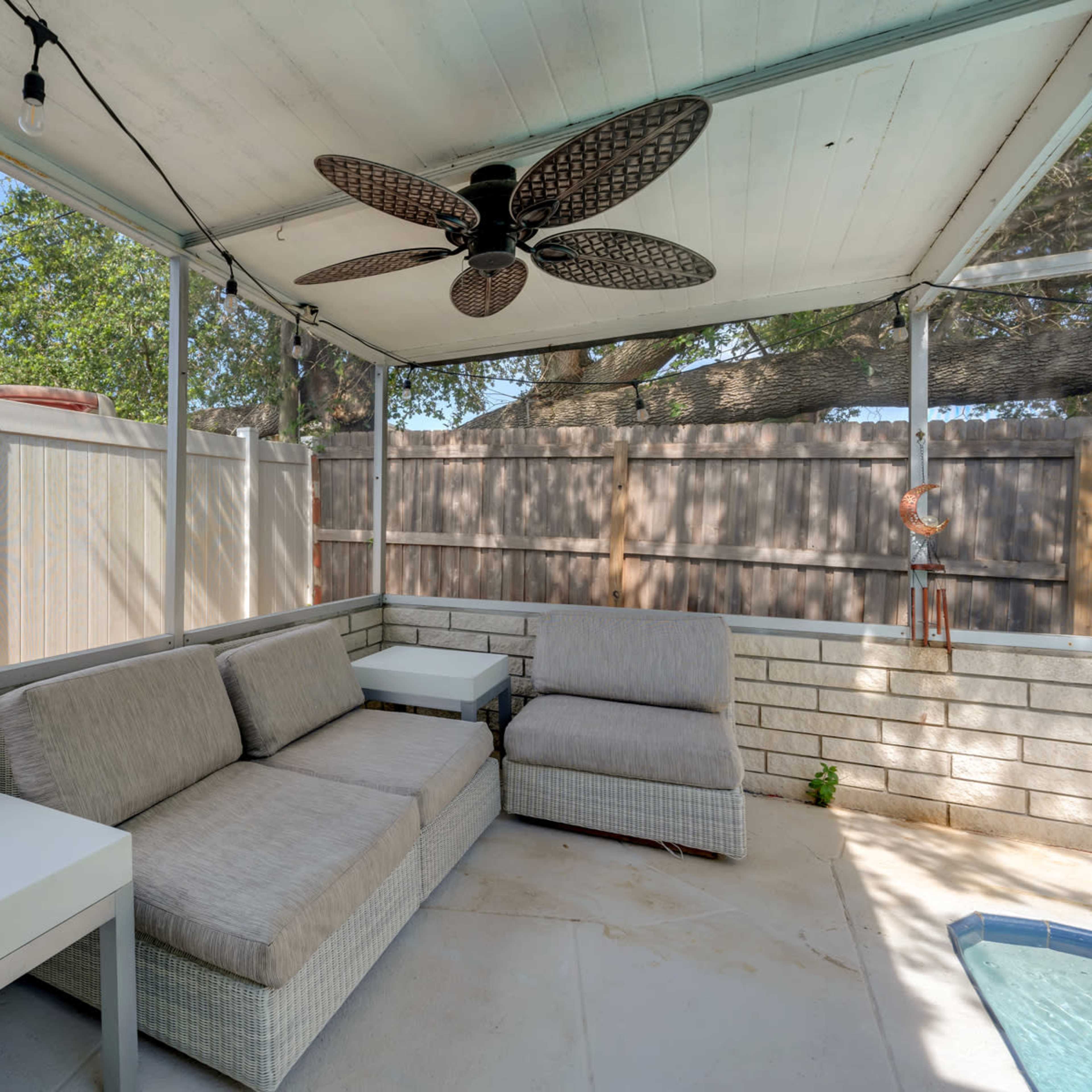 A covered patio area with outdoor seating and a ceiling fan, surrounded by a fence.