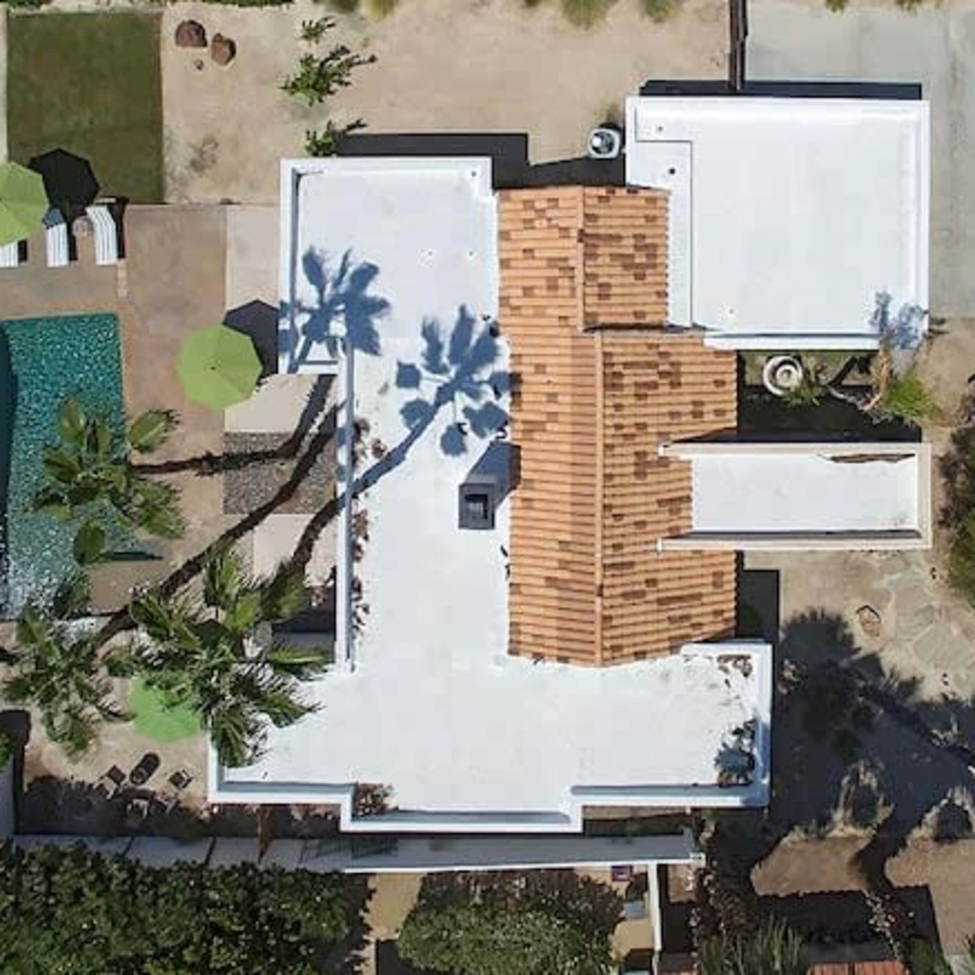 An aerial view of a modern residential property features a white-roofed house, a rectangular swimming pool, and several palm trees.