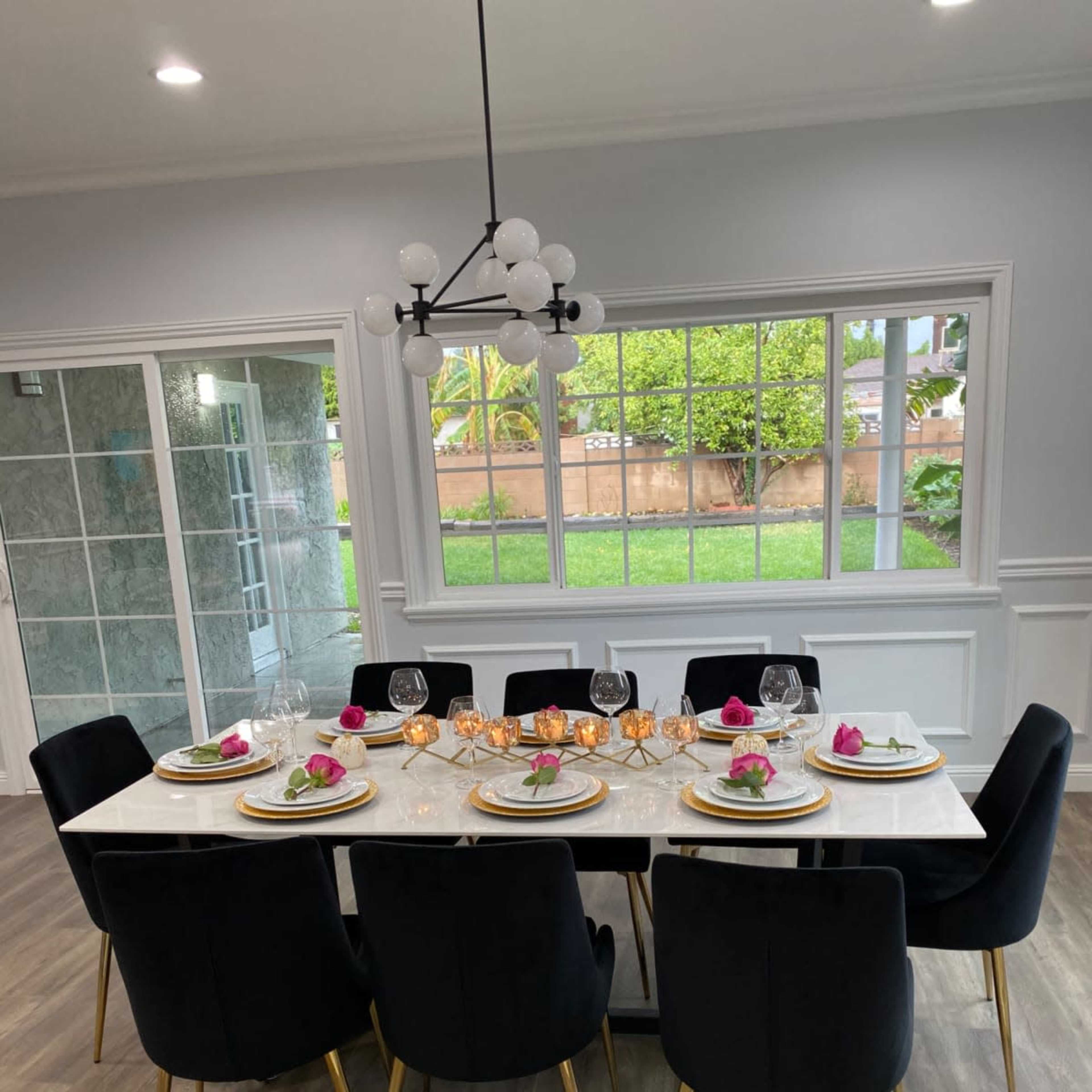 A dining table is set for a formal meal, featuring elegant dishware and floral centerpieces, in a well-lit room with large windows.