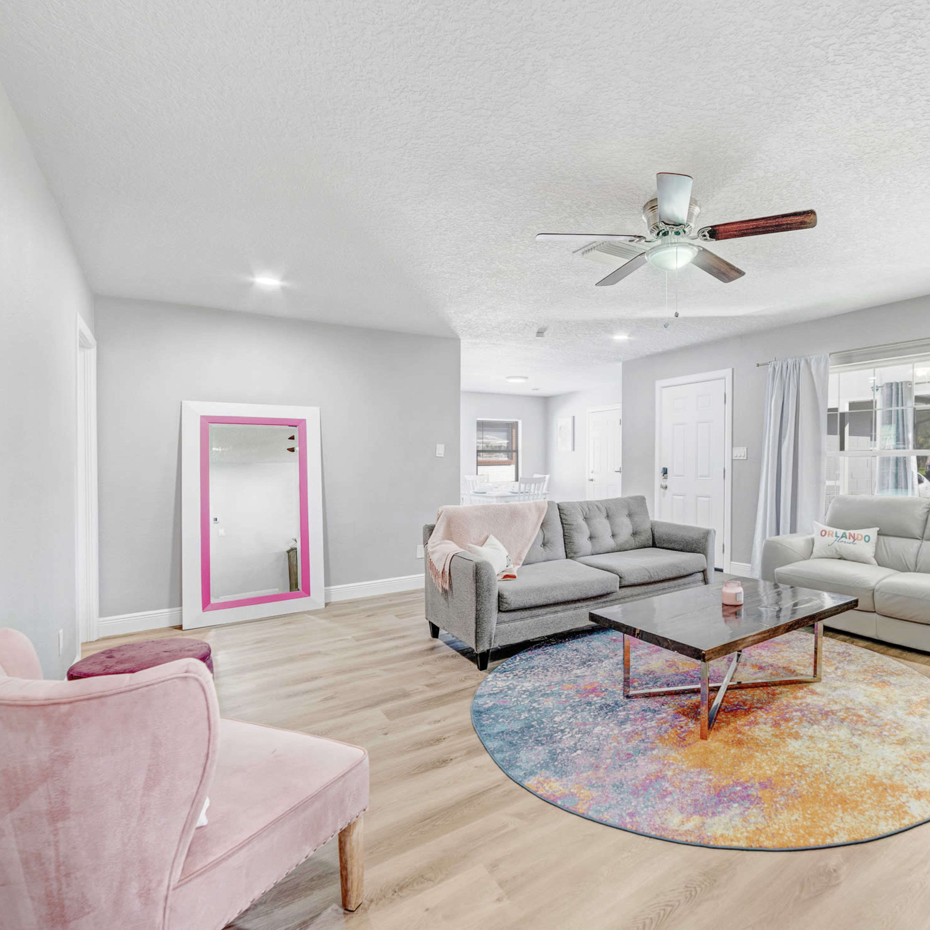 The living room features a light-colored sofa, a gray sectional, a circular rug, and a large mirror with a pink frame against the wall.