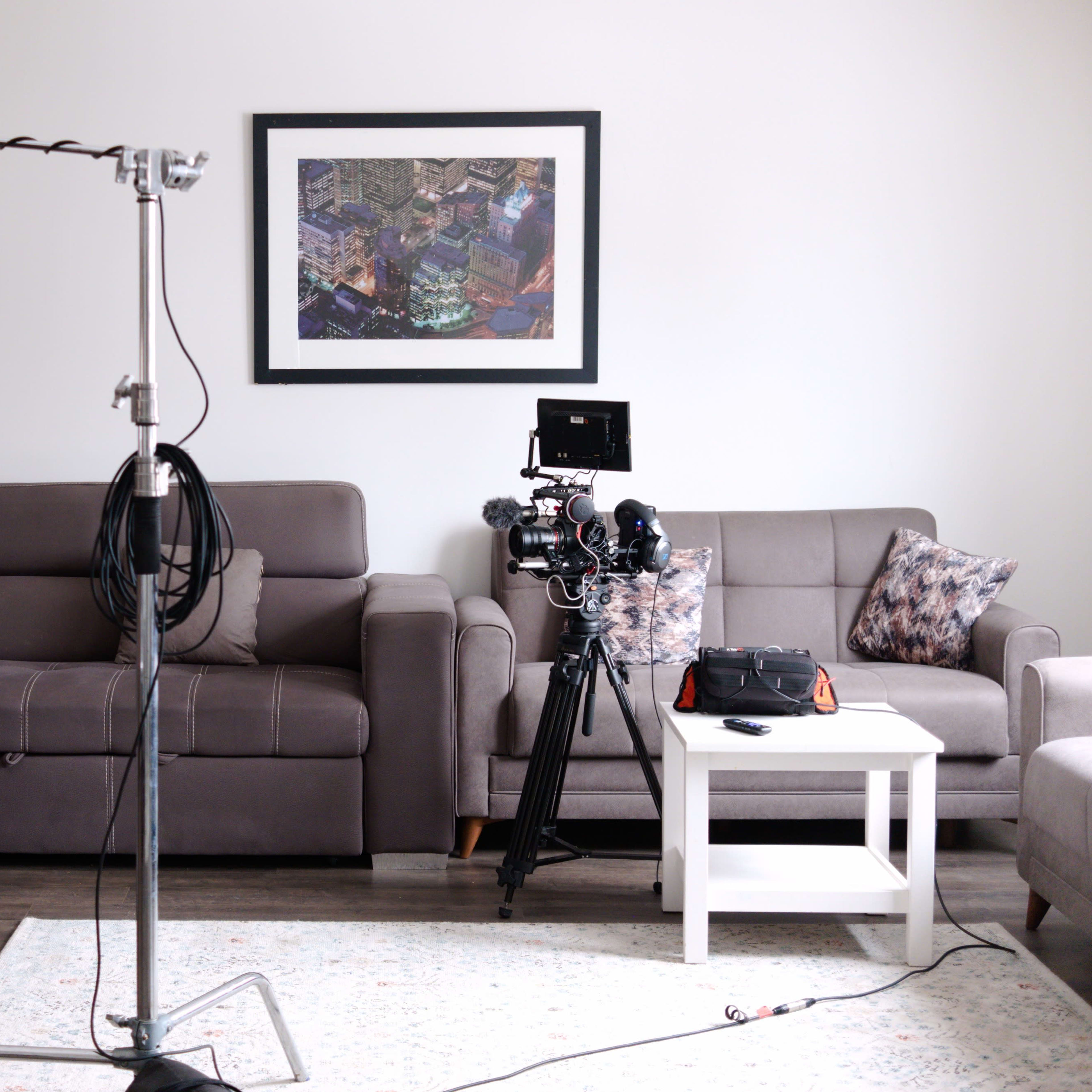 A living room is set up for a film shoot with a camera on a tripod, a microphone on a boom stand, and two couches arranged around a coffee table.