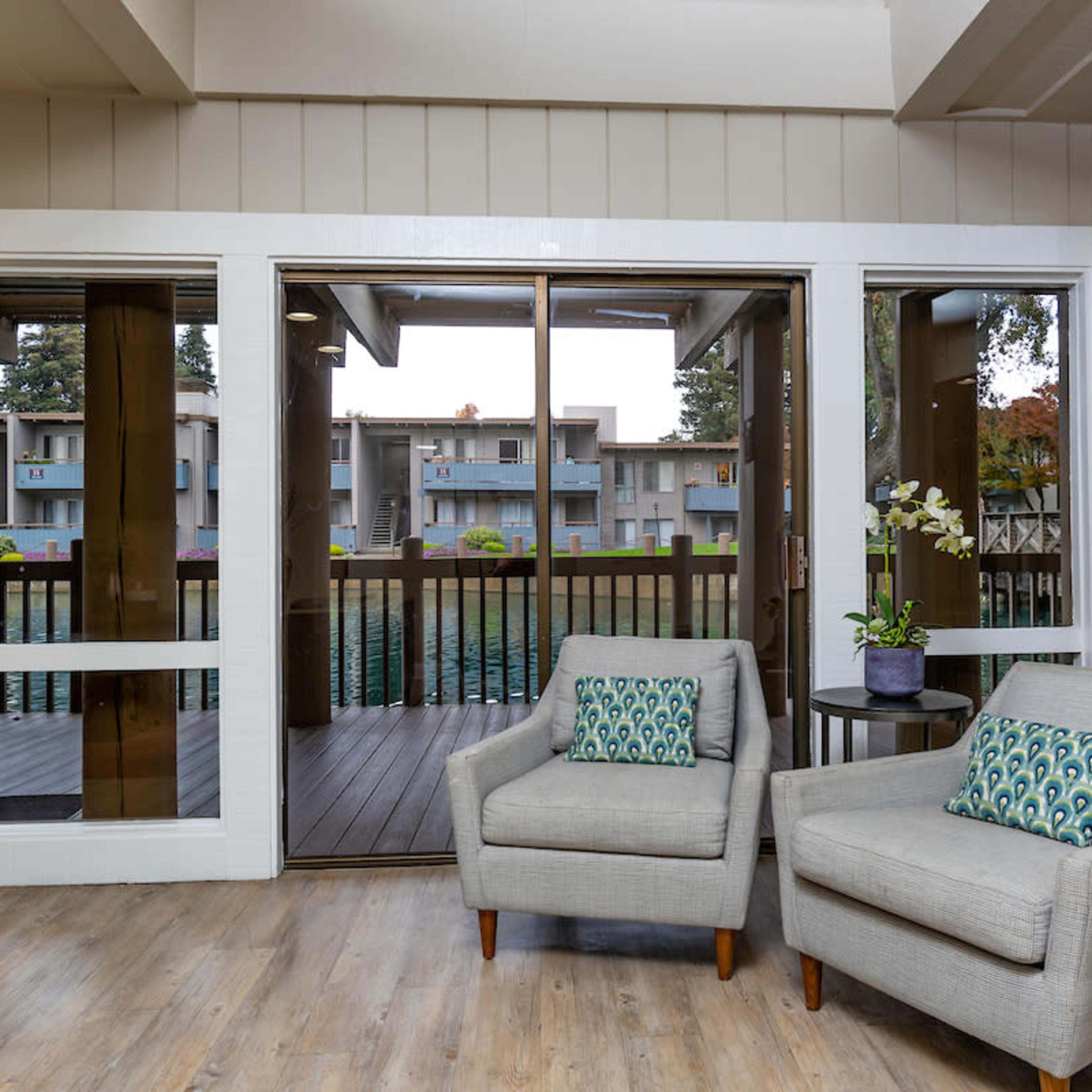 A pair of gray armchairs with decorative cushions are positioned beside a door that opens onto a wooden deck overlooking a small body of water.