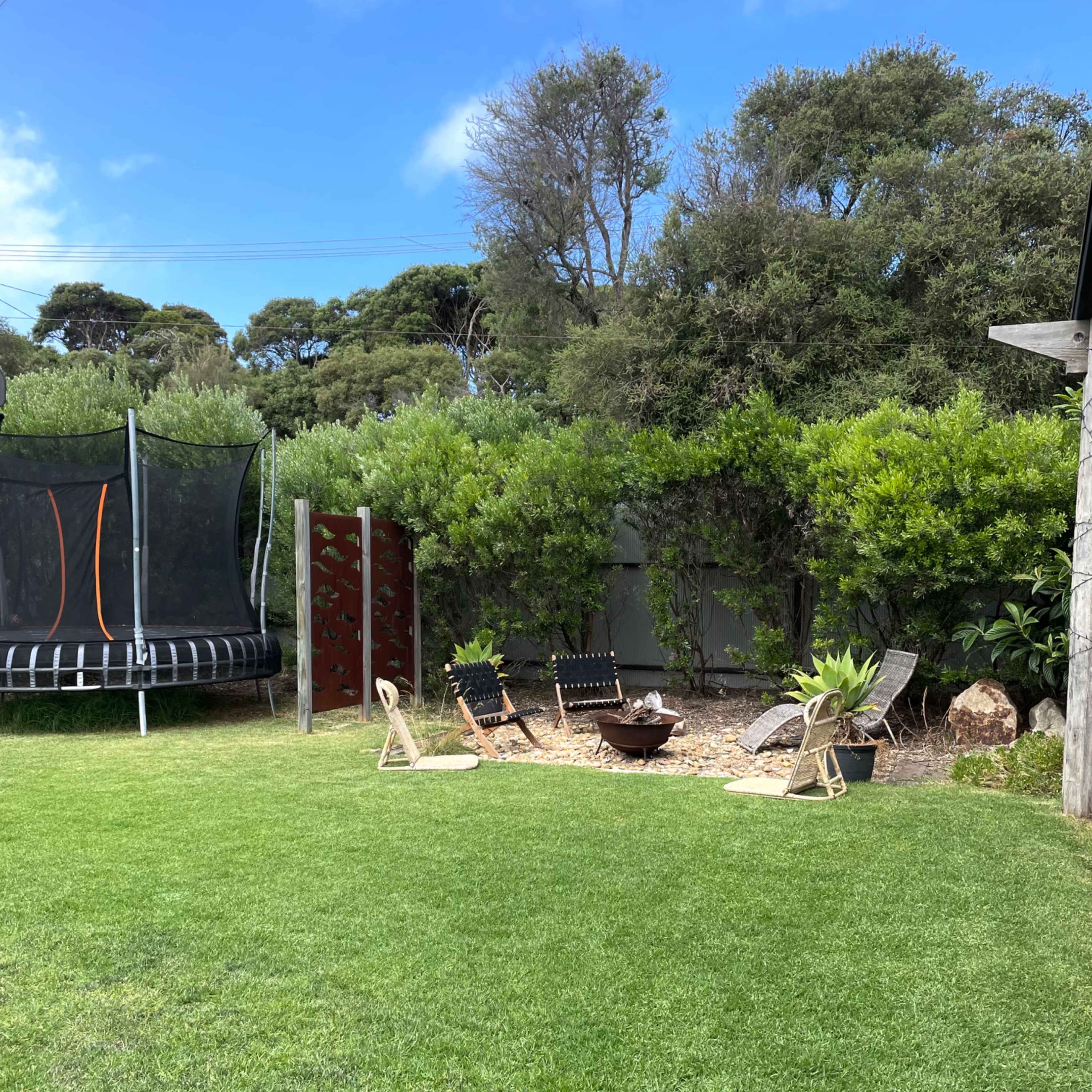 A grassy backyard featuring a trampoline, seating around a fire pit, and lush greenery in the background.