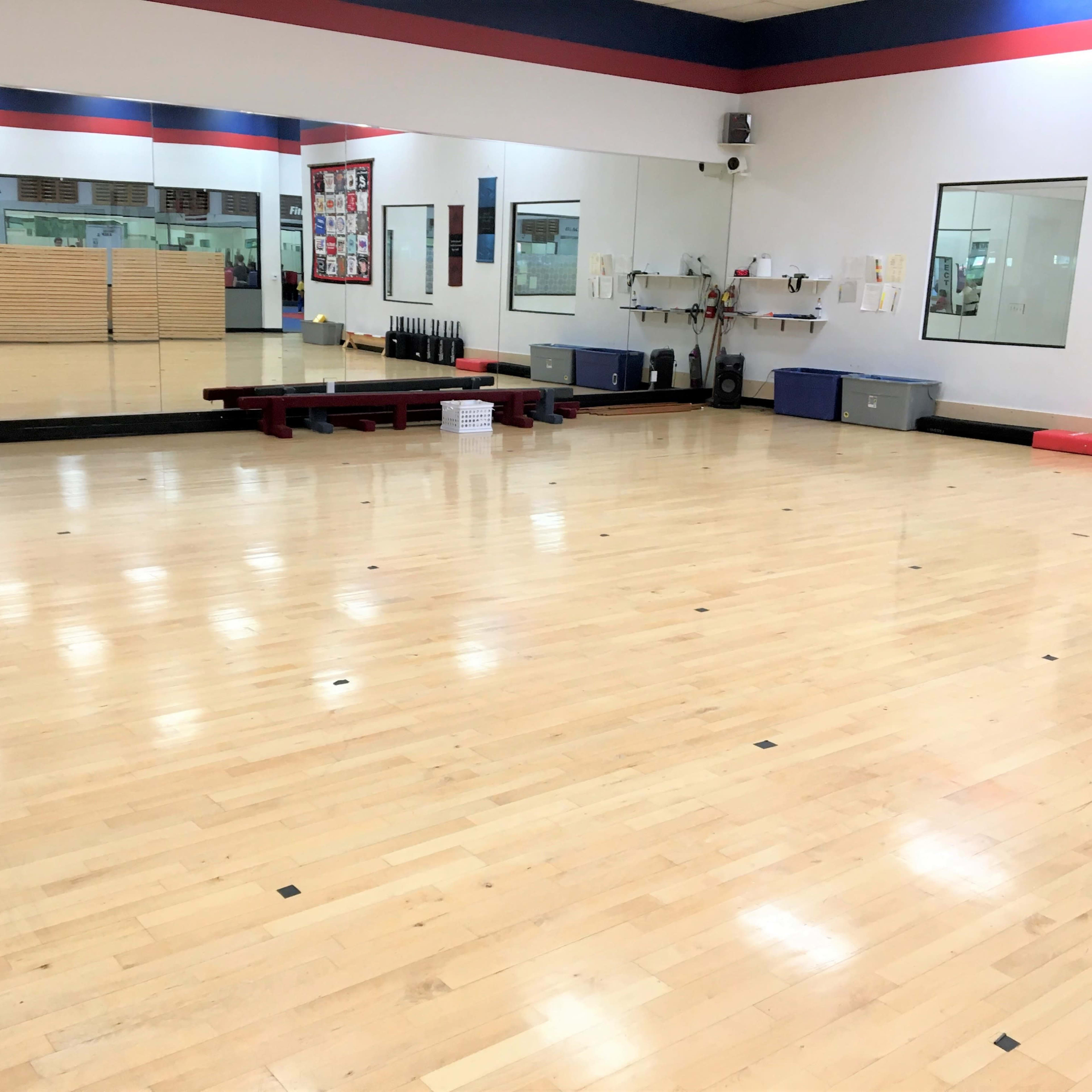 A spacious dance or exercise studio with wooden flooring, mirrors on one wall, and various fitness equipment stored on the side.