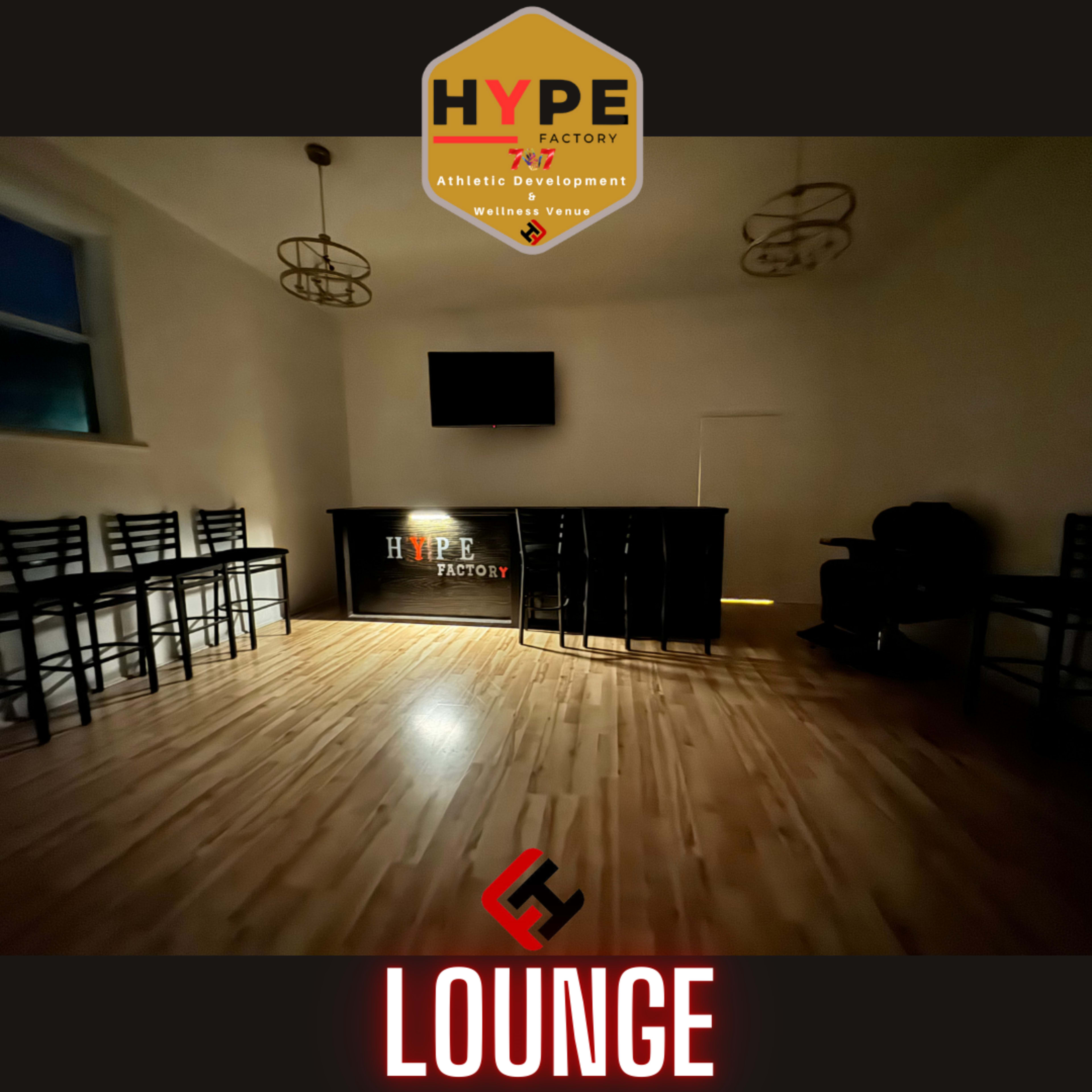 A dimly lit lounge area with wooden flooring, a bar counter adorned with the "HYPE FACTORY" logo, and a few high chairs.