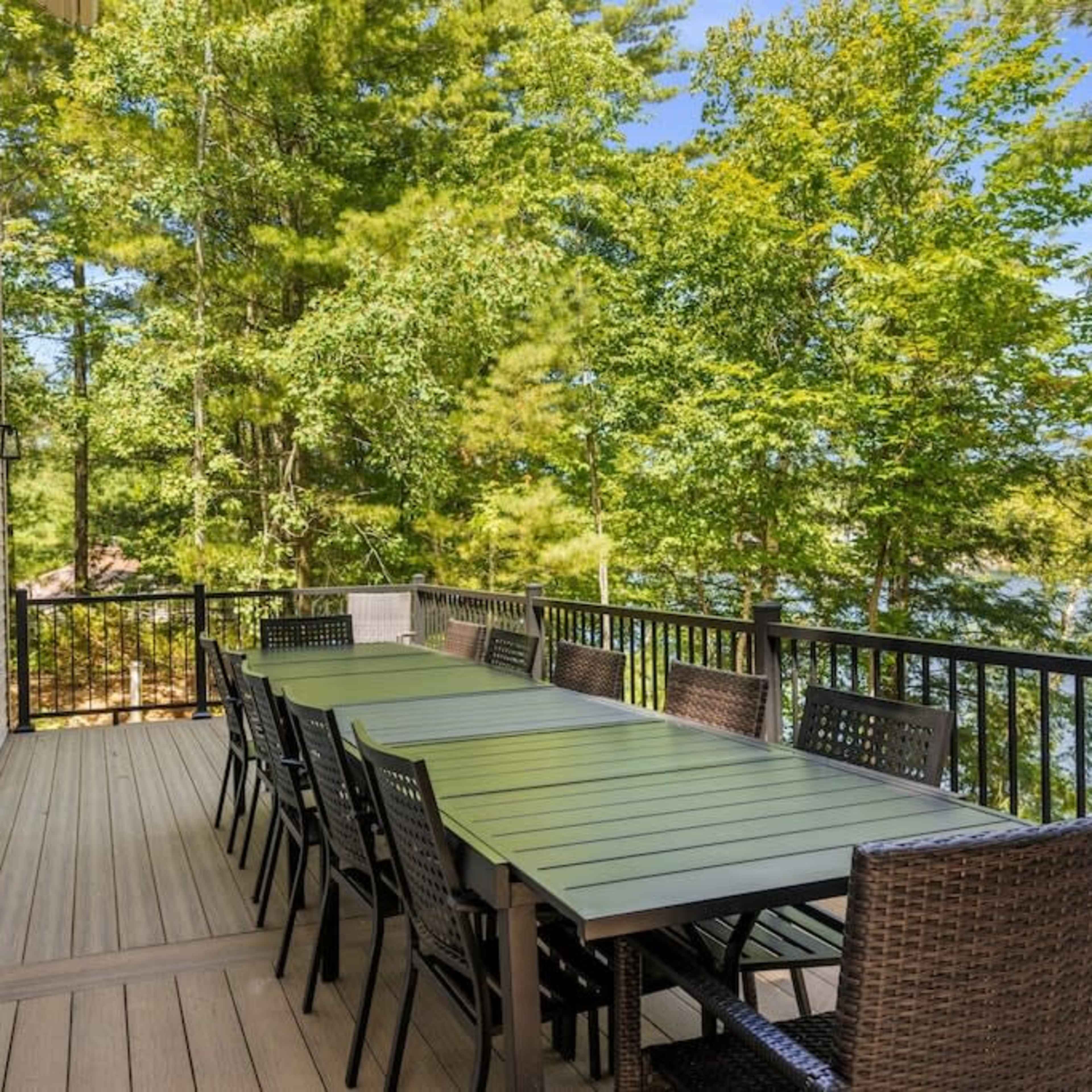 A long, rectangular dining table with multiple chairs is set on a wooden deck overlooking a body of water and surrounded by trees.