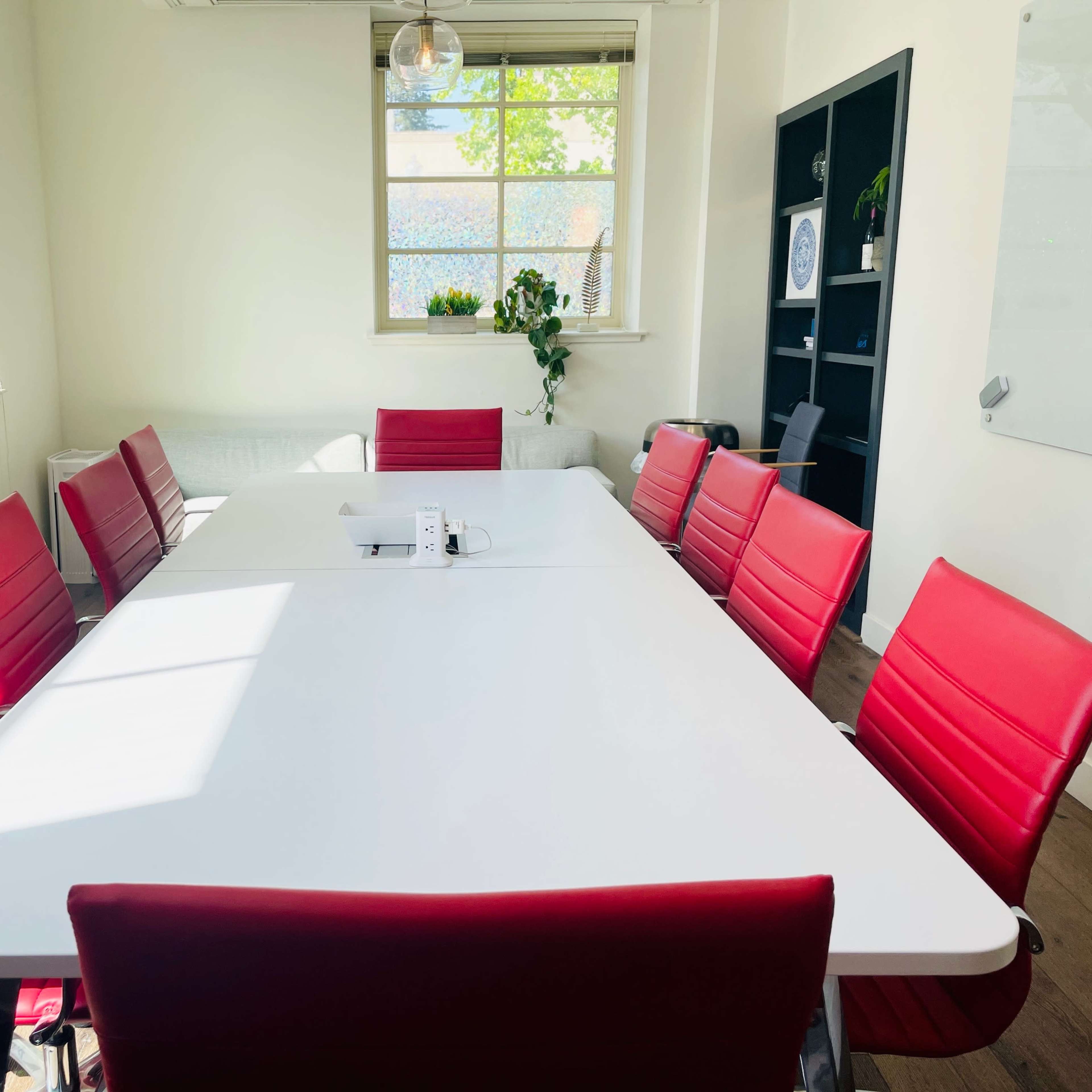 A bright meeting room features a large white table surrounded by red chairs, with sunlight streaming through a window.