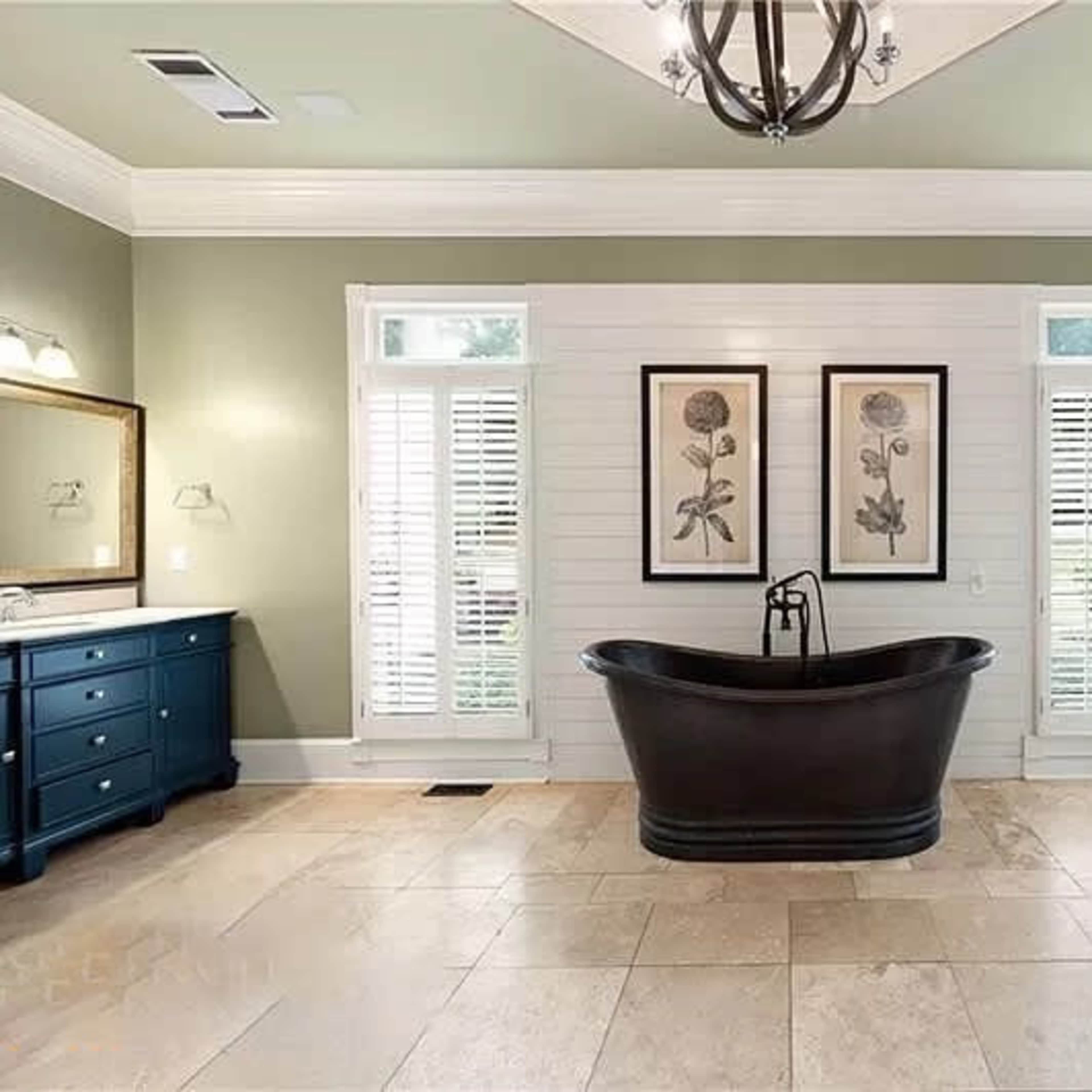 A spacious bathroom featuring a freestanding black tub centered between two vanities with dark blue cabinets, framed botanical prints on the wall, and light-colored tiled flooring.