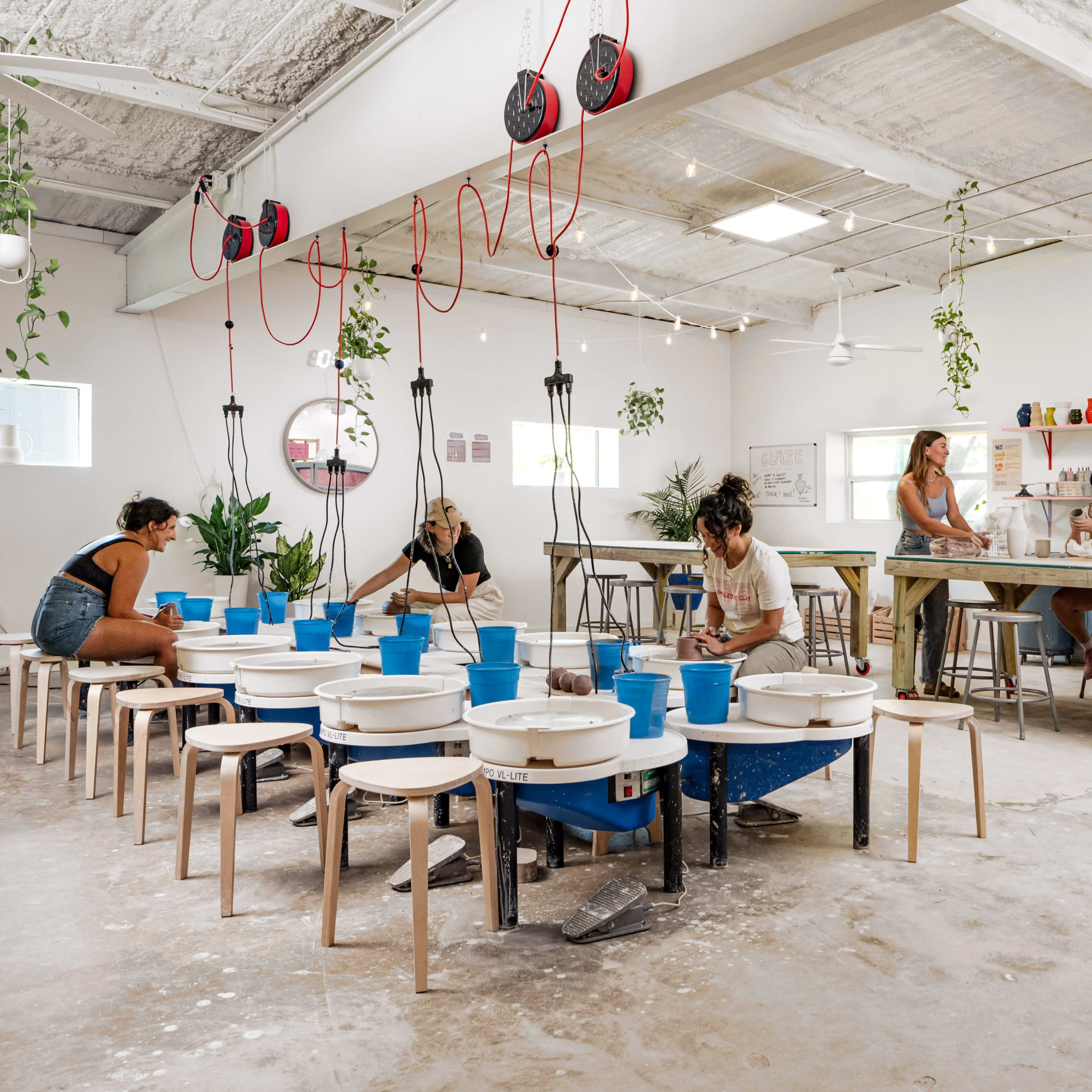 A spacious pottery studio features several pottery wheels arranged in a semicircle, with individuals working on projects and a communal area with a countertop and tools in the background.