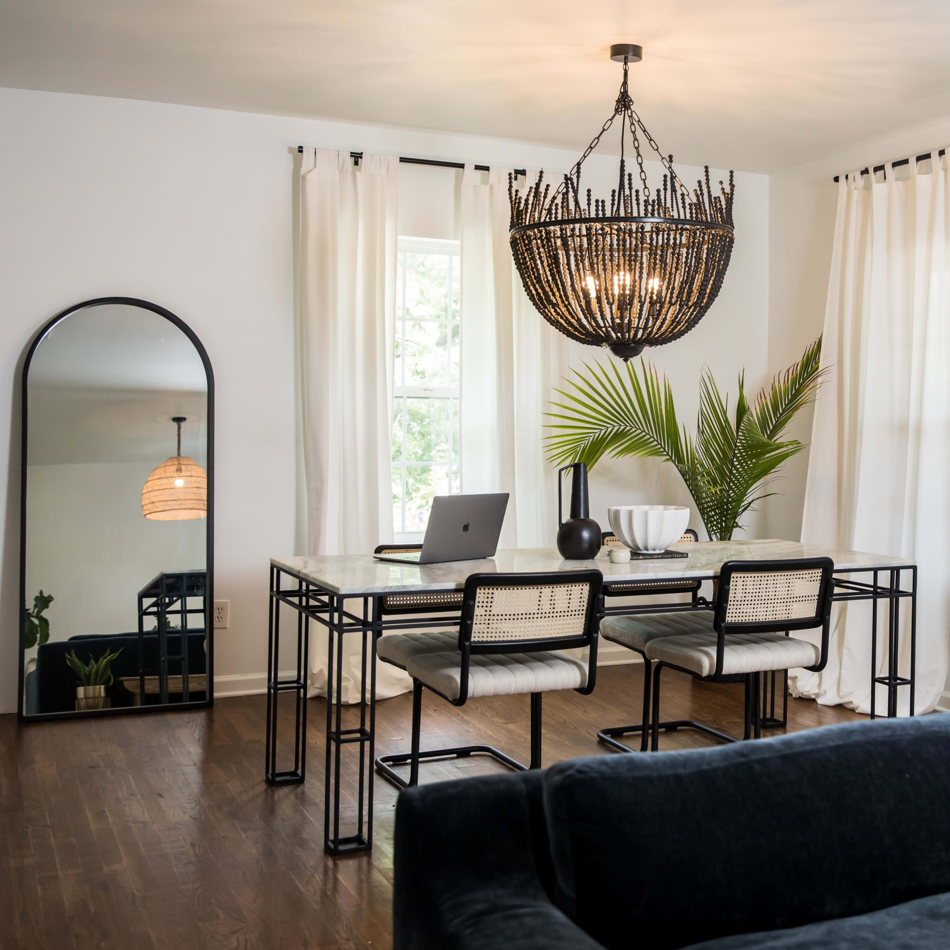 A dining area features a marble table with four chairs, a large mirror, and a stylish chandelier, surrounded by bright curtains and a potted plant.