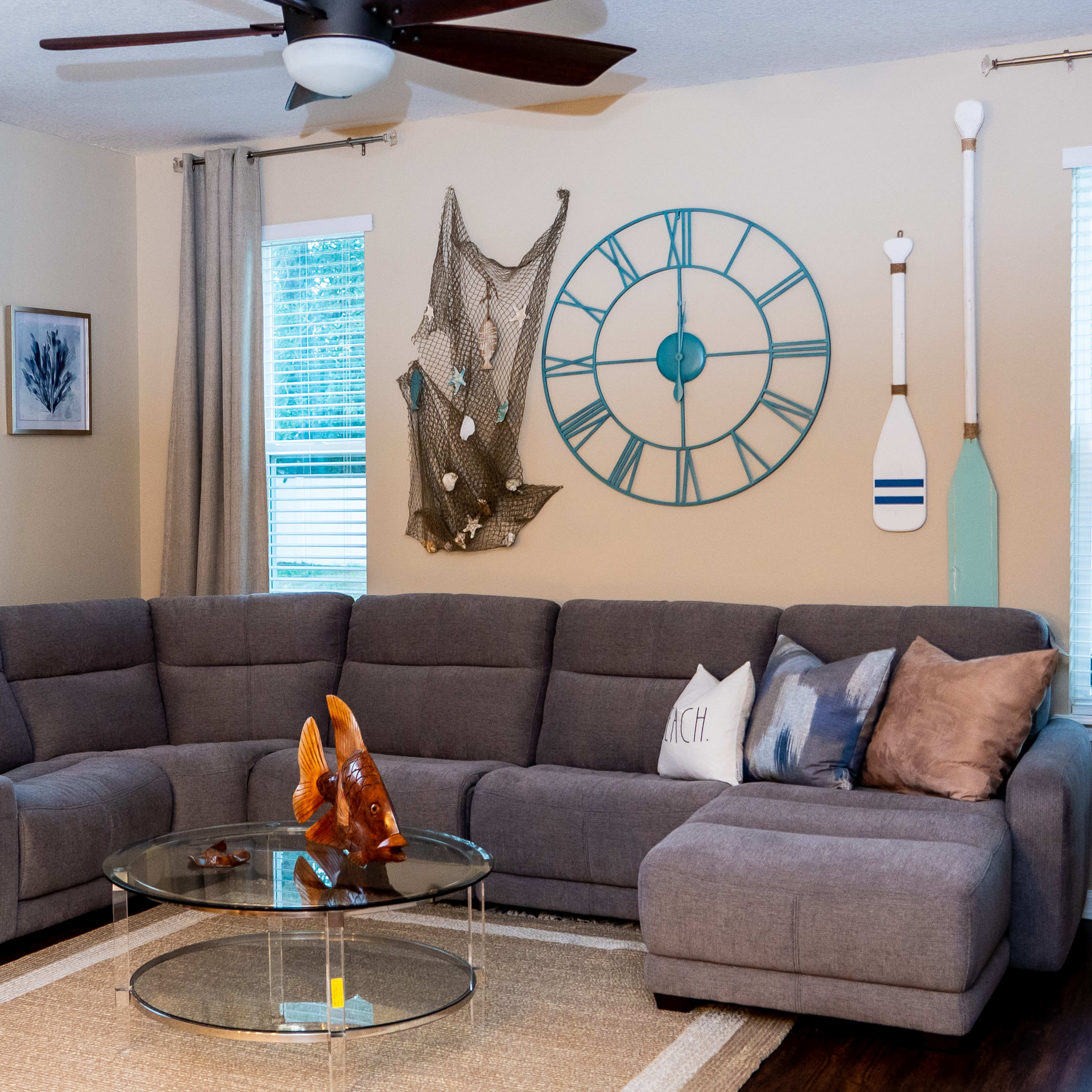 A gray sectional sofa with decorative pillows is arranged around a glass coffee table in a brightly lit living room featuring nautical-themed decorations on the walls.