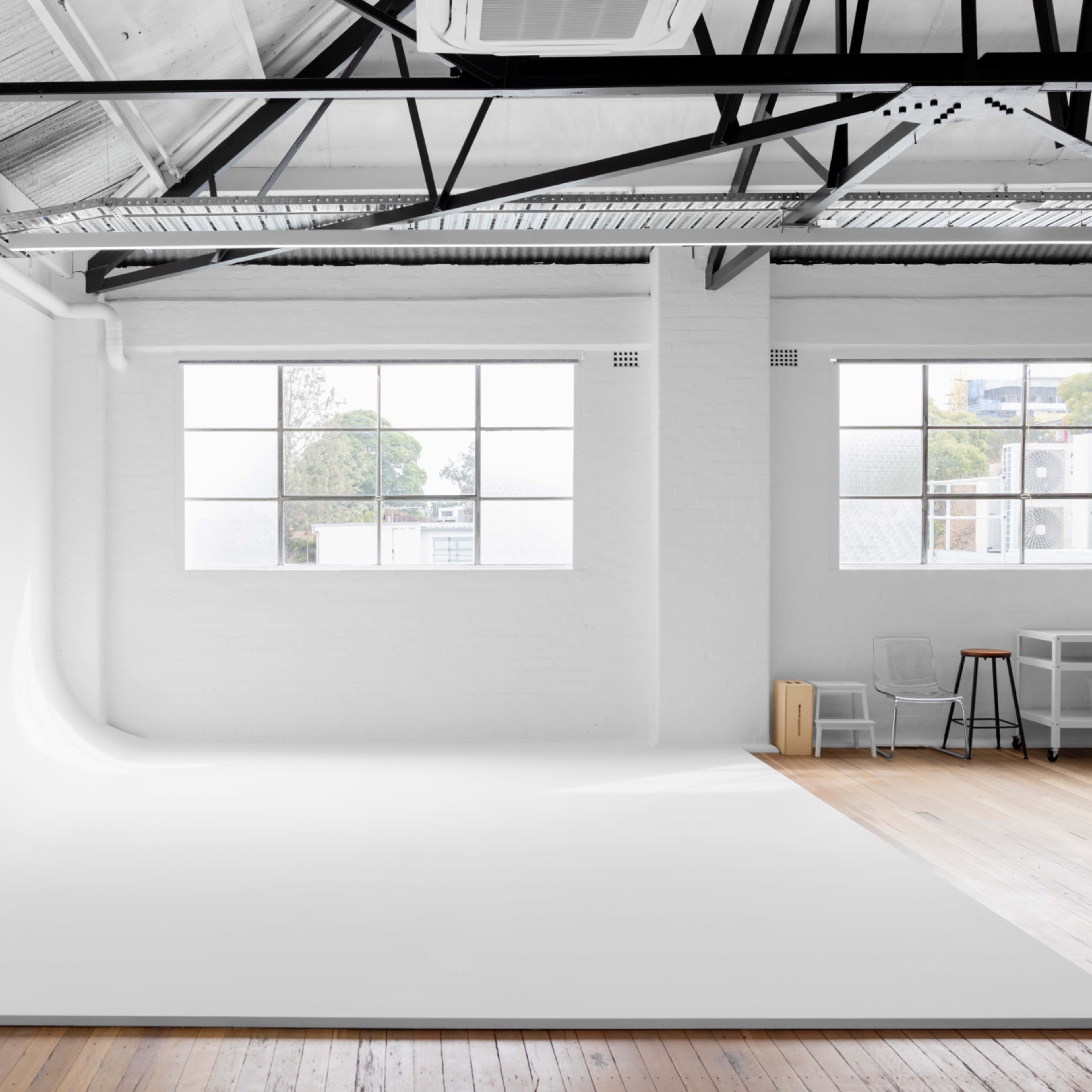 A spacious photography studio with white walls, a seamless backdrop, large windows, and wooden flooring.