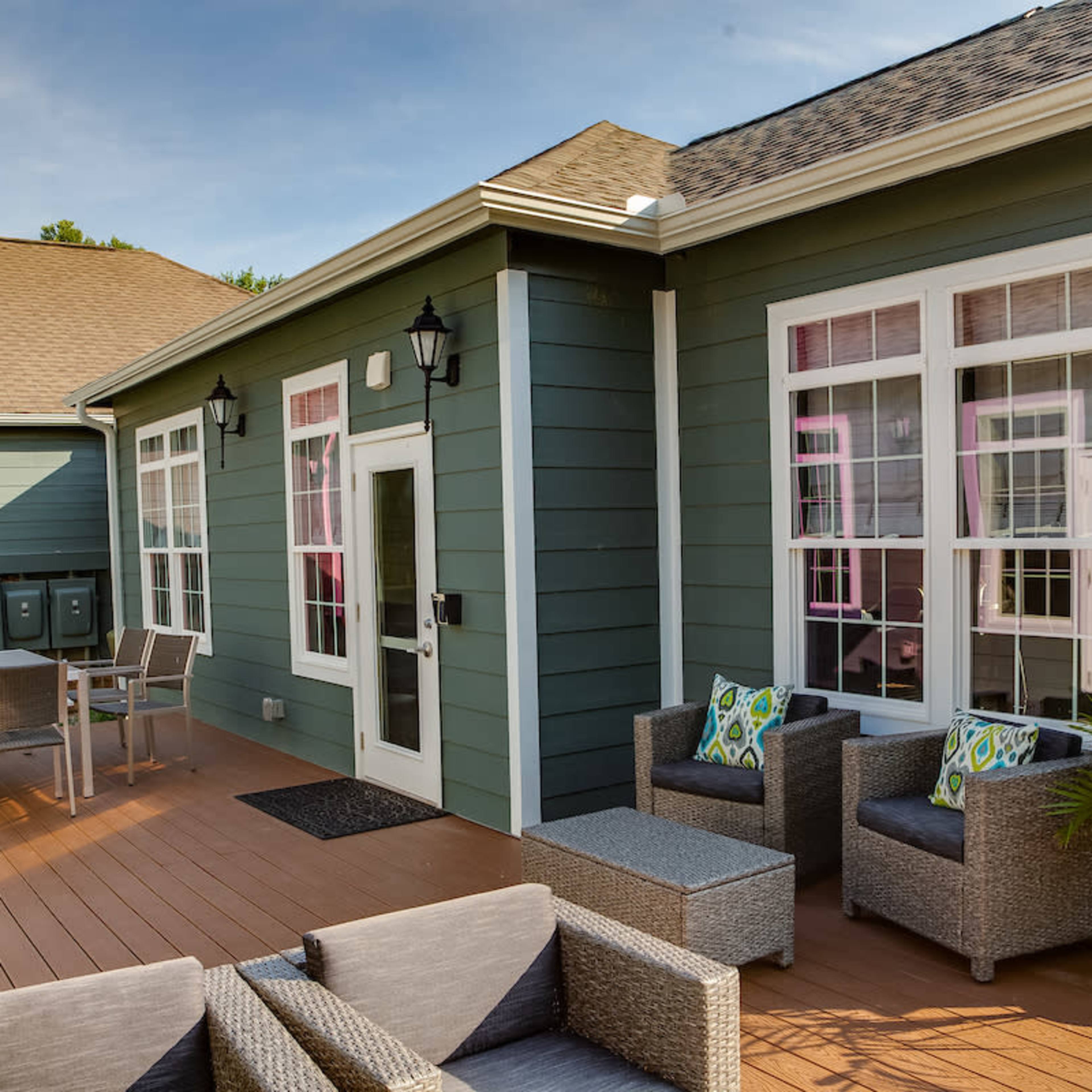 A well-lit outdoor patio area with grey wicker furniture, a dining table, and green walls on a house.