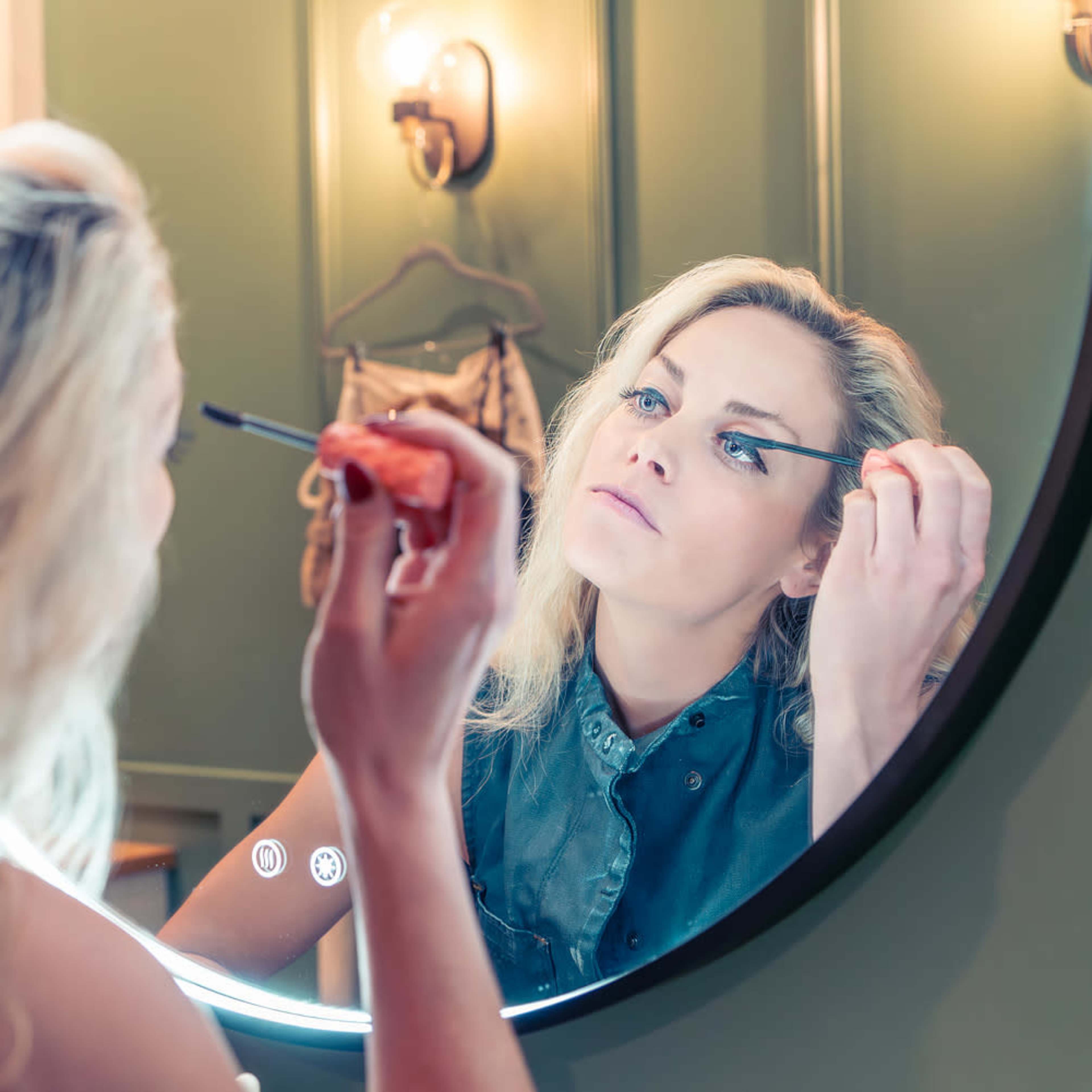 A woman applies mascara while looking into a circular mirror.