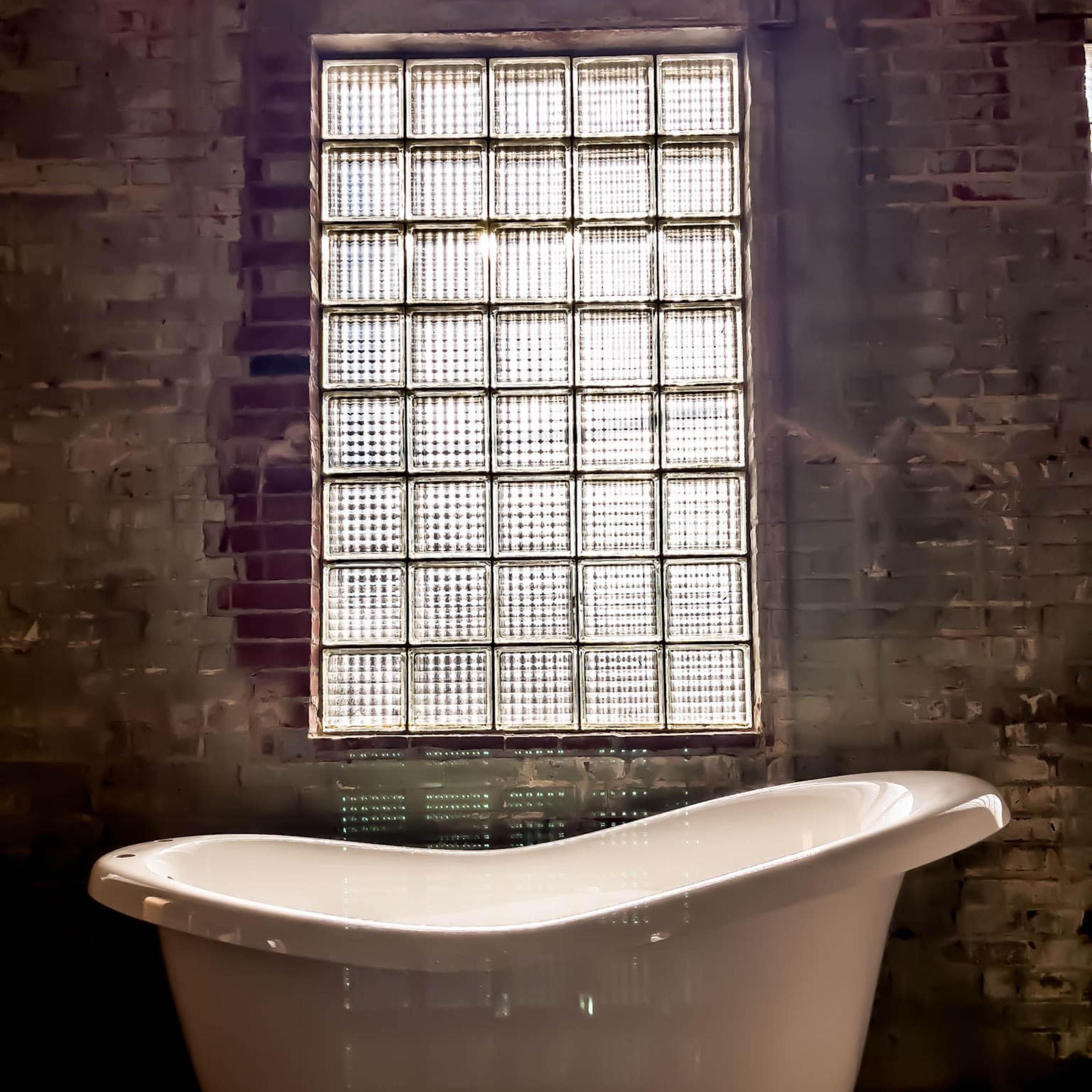 A white freestanding bathtub is positioned in front of a large glass block window set into a brick wall.
