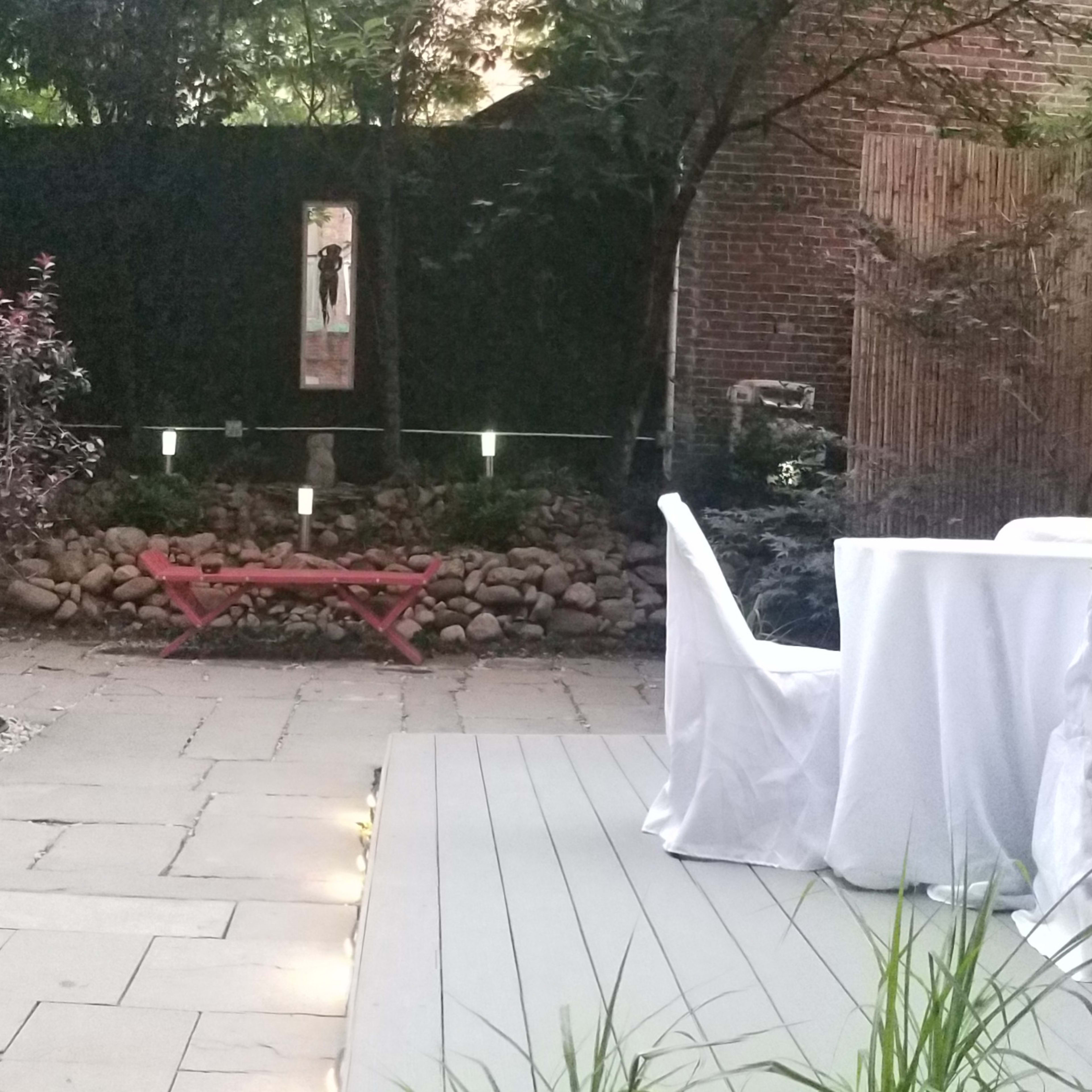 A small outdoor garden area features a stone path, a wooden bench, and a table covered with a white tablecloth, surrounded by greenery and decorative rocks.