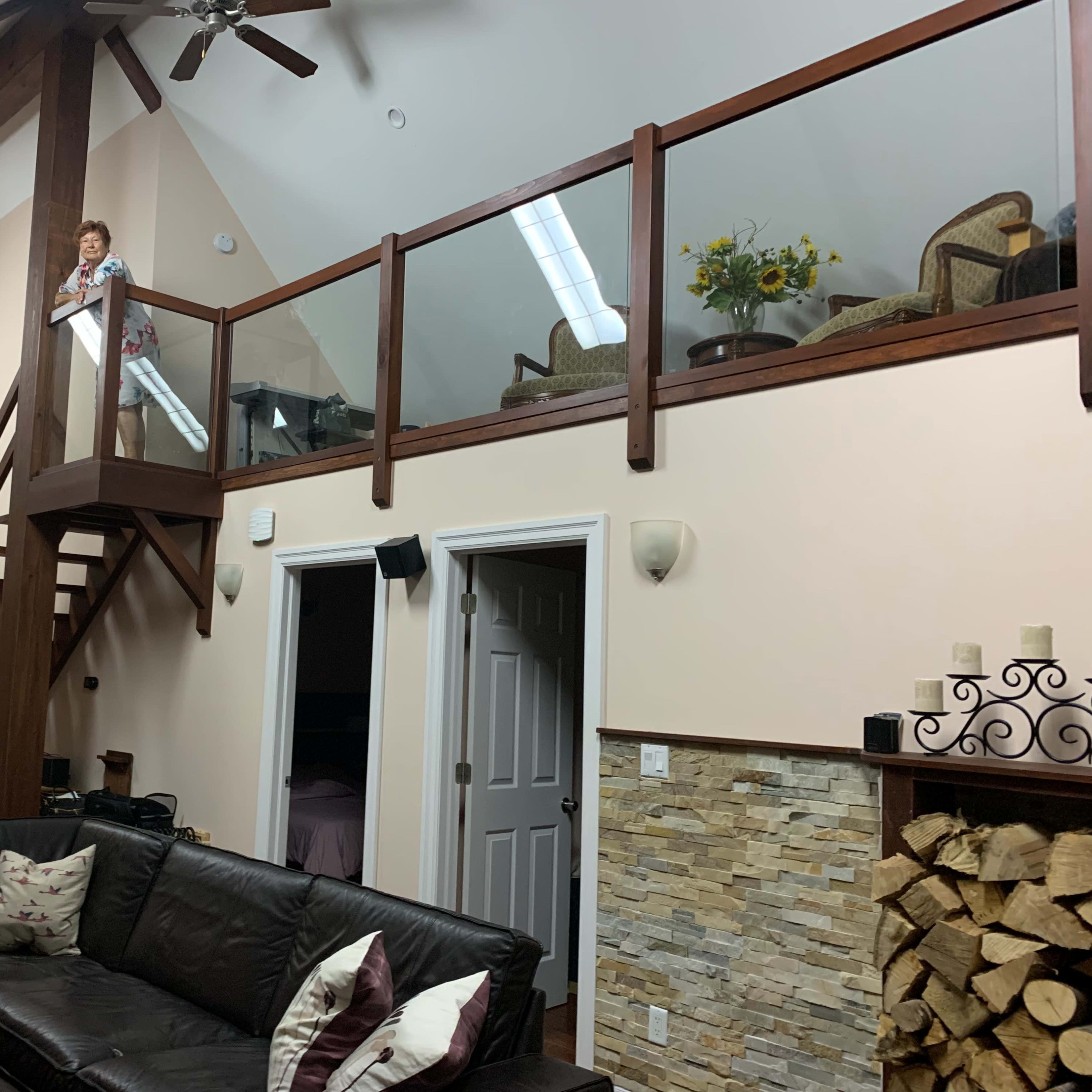 A woman stands on a wooden loft balcony overlooking a living room with a large sectional sofa and a stone fireplace, while stairs lead down to the dining area.