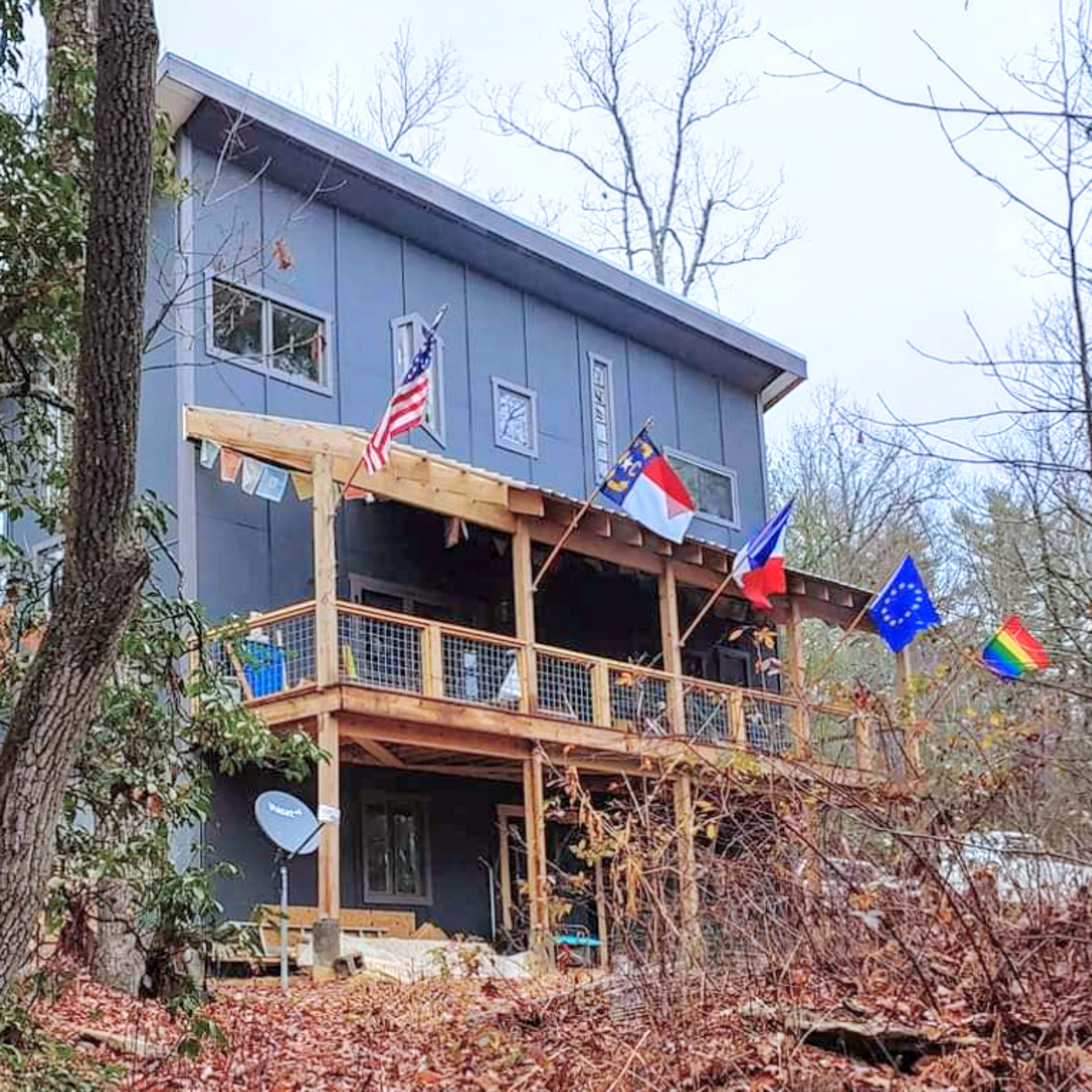 A three-story house with a wooden deck displays multiple flags, including the American flag, a state flag, and an EU flag, surrounded by autumn foliage.