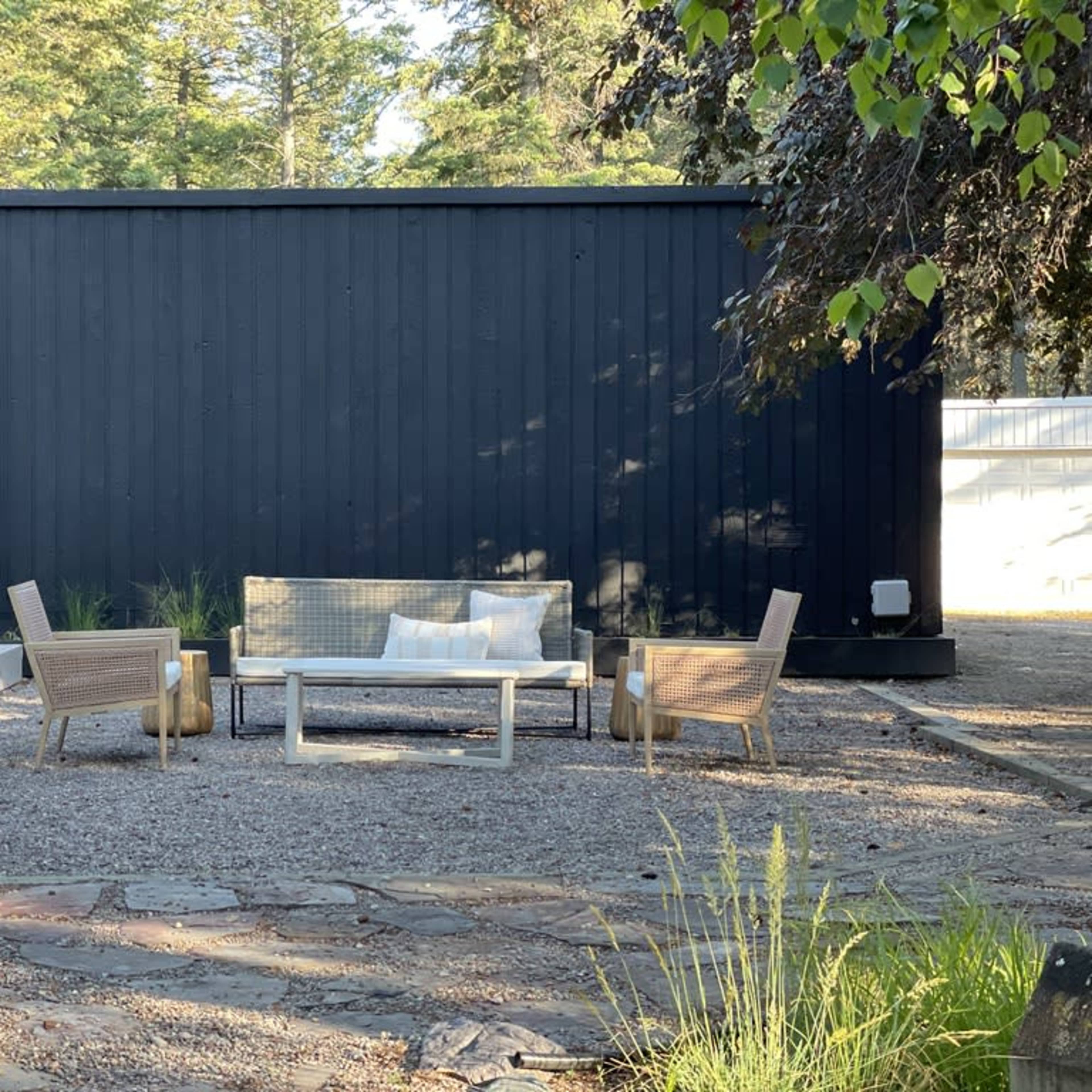 A modern outdoor seating area with a rectangular table and four chairs placed on gravel in front of a black wooden wall.