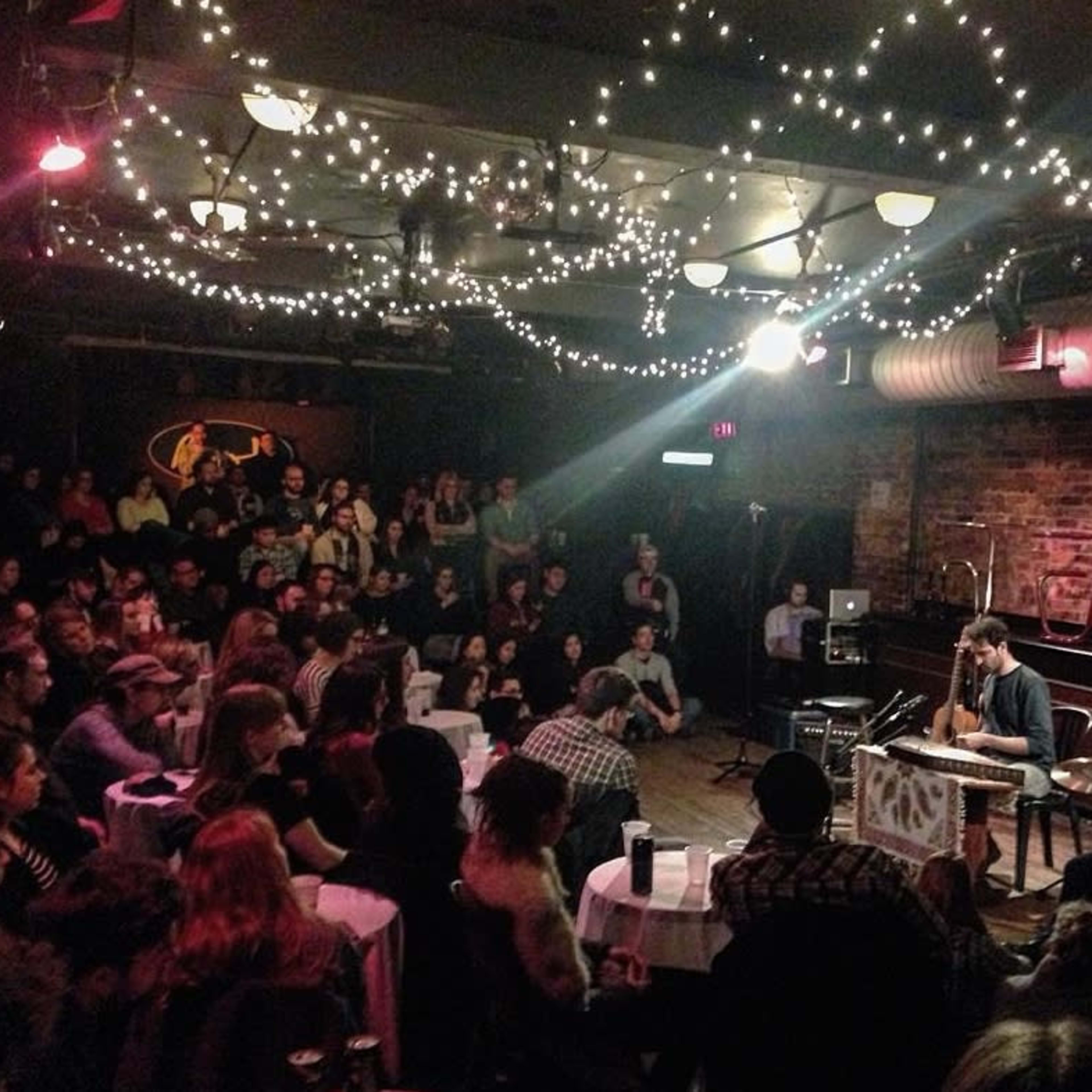 A musician performs on stage in a crowded venue illuminated by string lights.