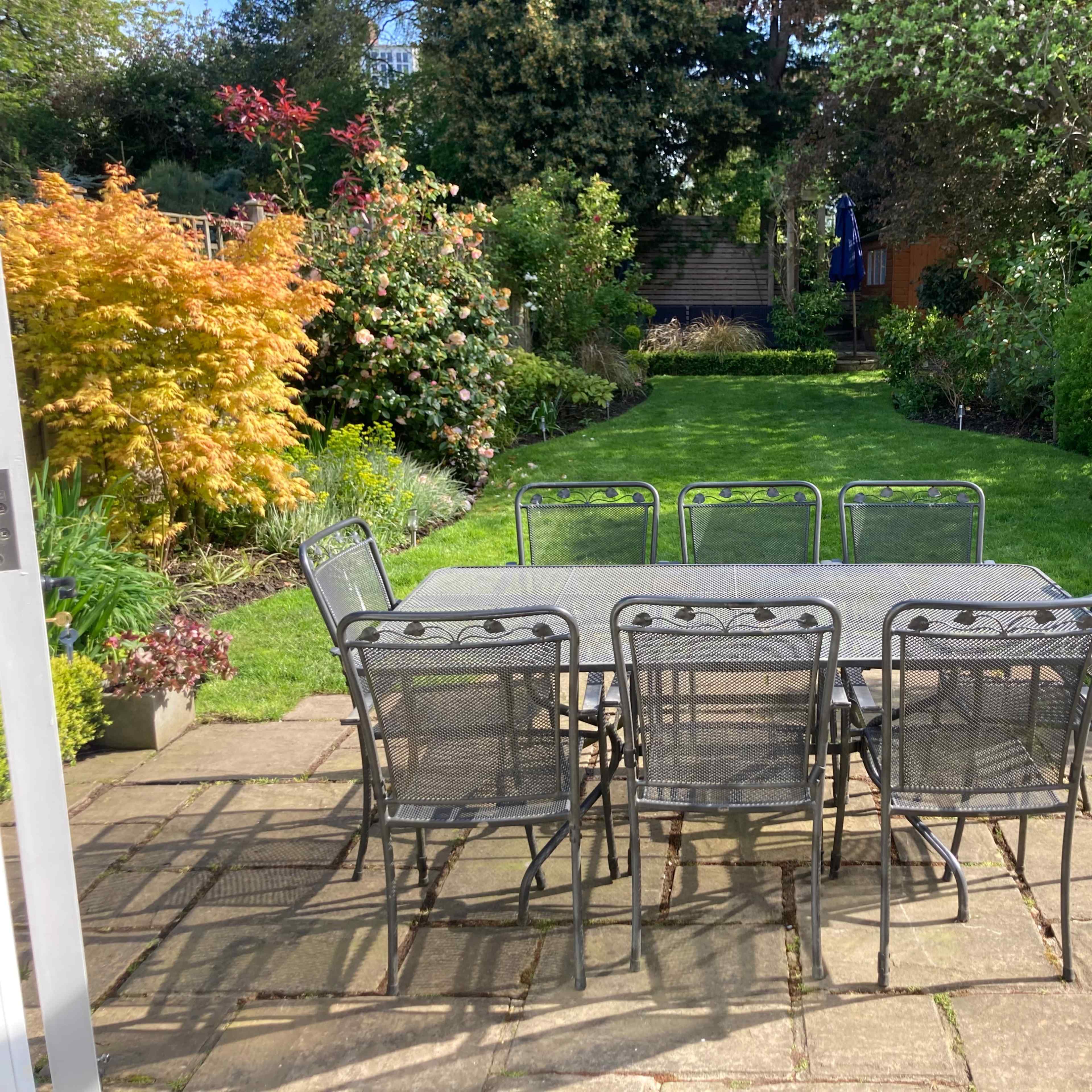 A patio table surrounded by eight chairs overlooks a well-maintained garden with vibrant greenery and flowering plants.