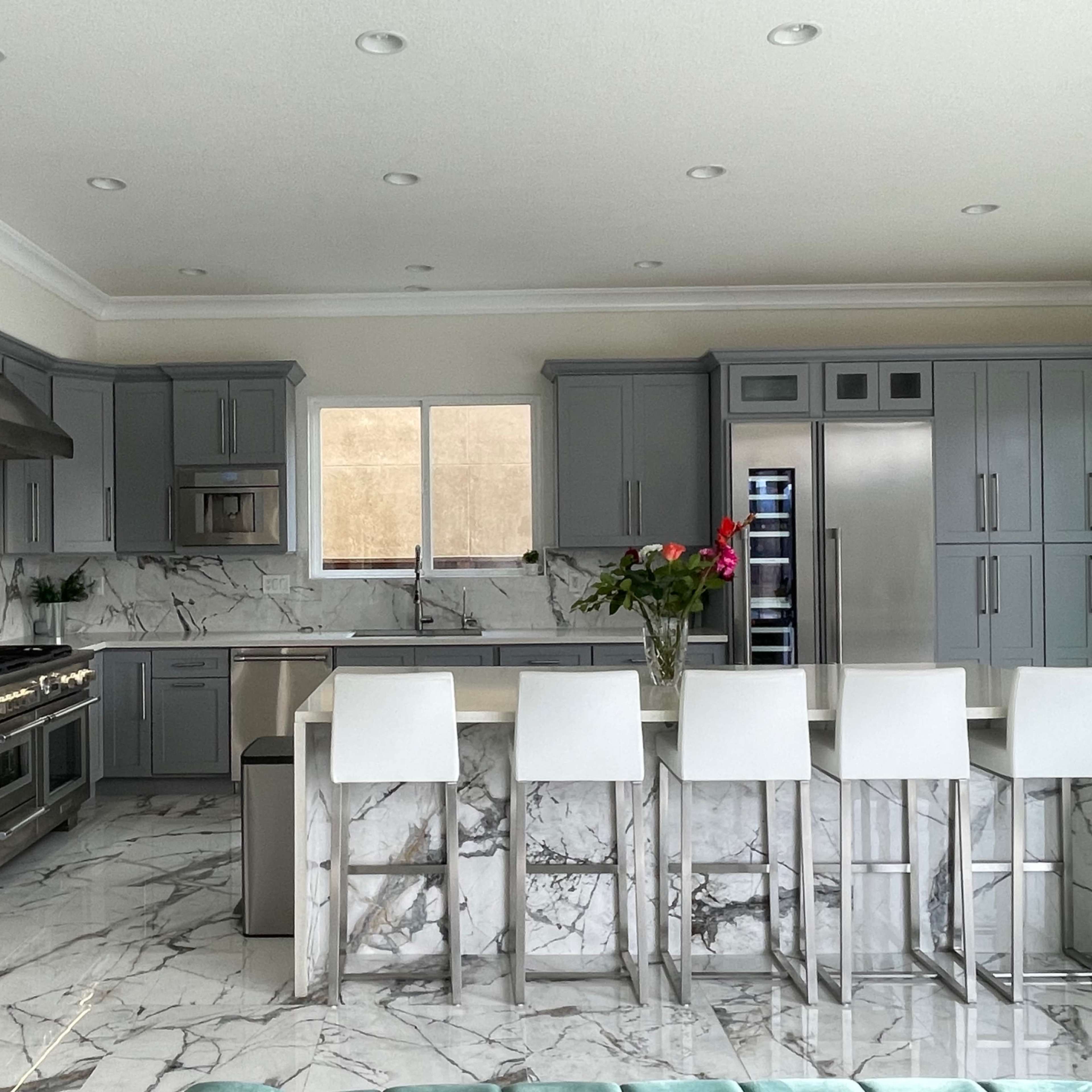 A modern kitchen features gray cabinets, marble countertops, and a large island with white stools.