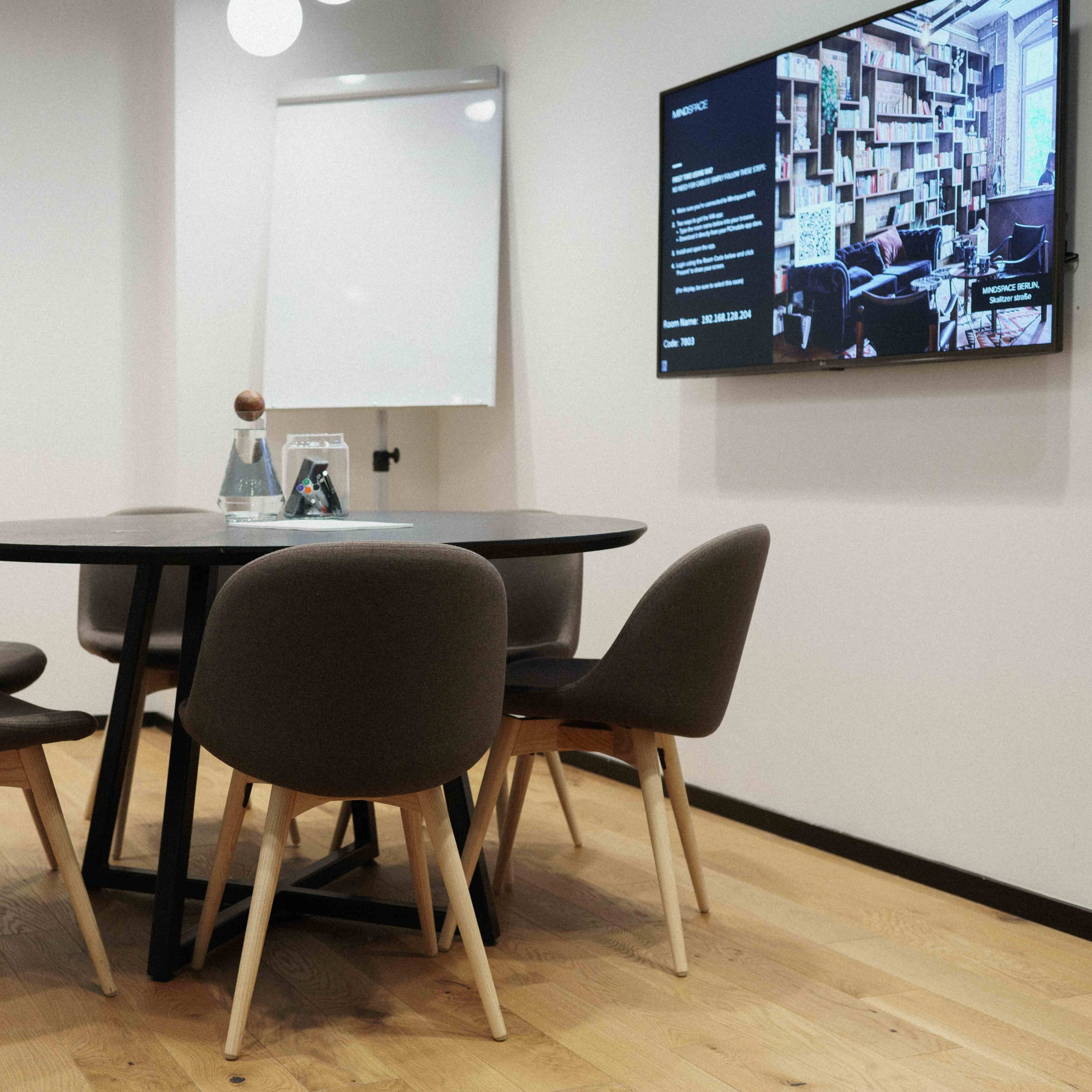 A modern conference room features a round table surrounded by five chairs, a whiteboard, and a wall-mounted display.