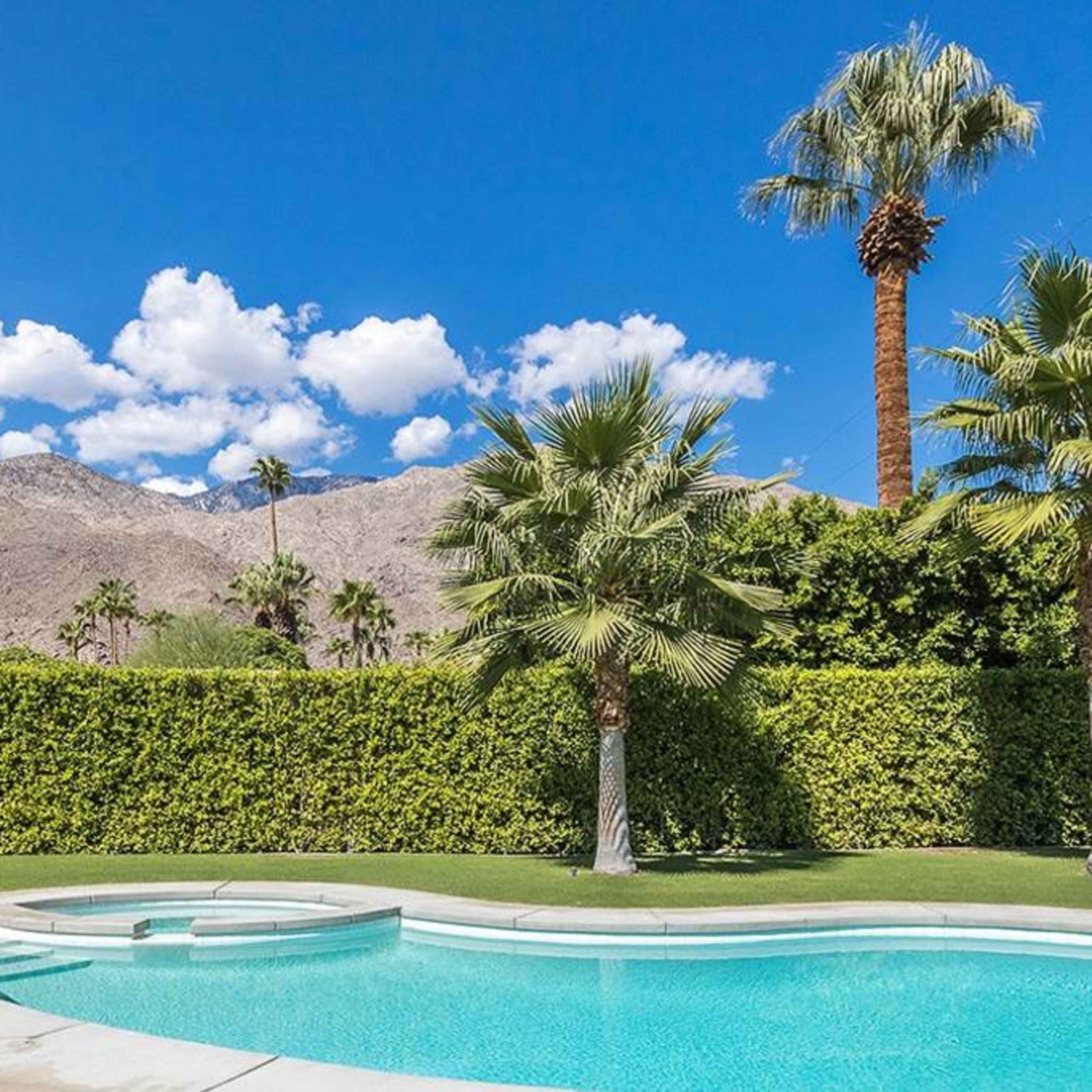 A swimming pool is set against a backdrop of mountains and palm trees under a blue sky with scattered clouds.