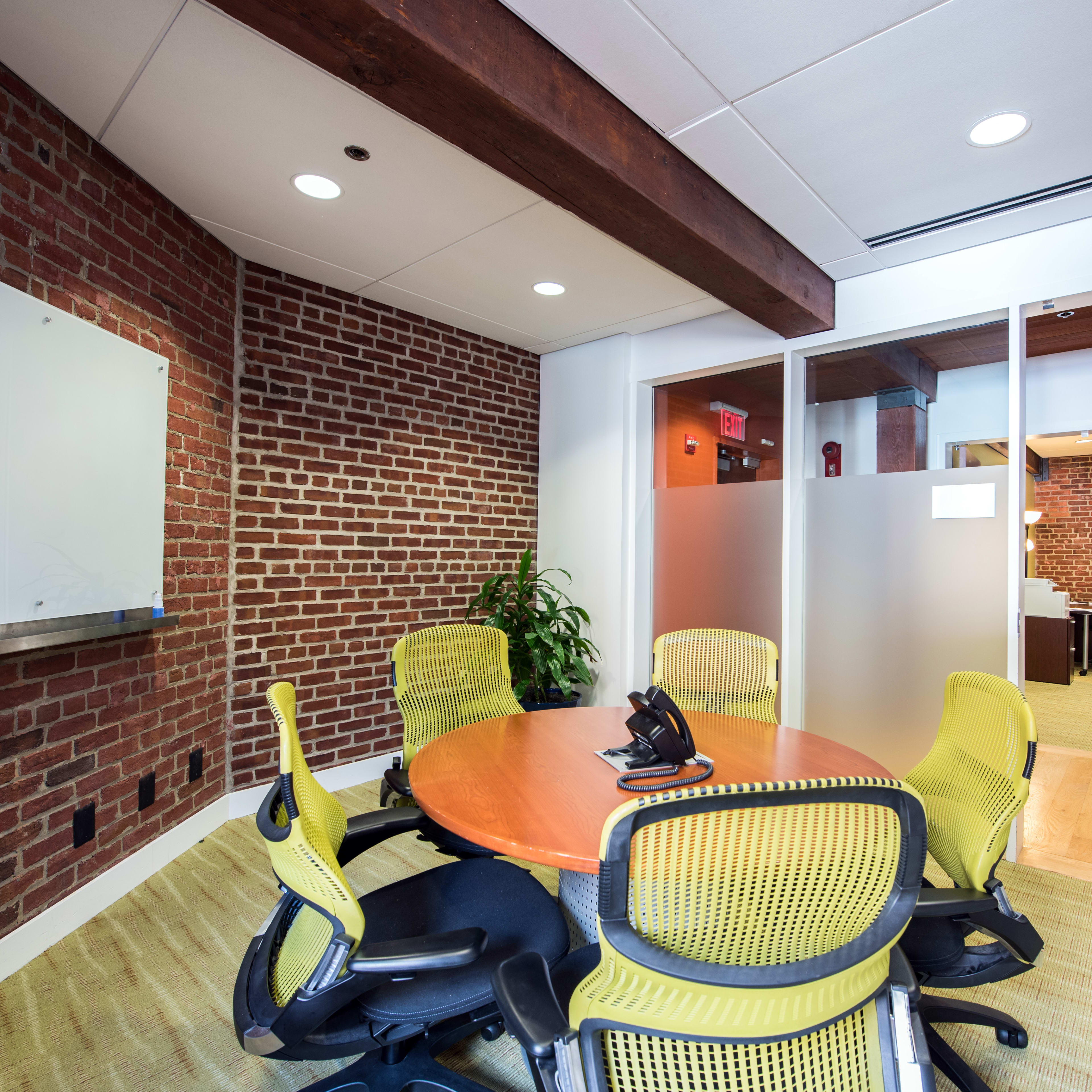 A conference room with a round wooden table surrounded by four yellow office chairs, set against a backdrop of exposed brick walls.