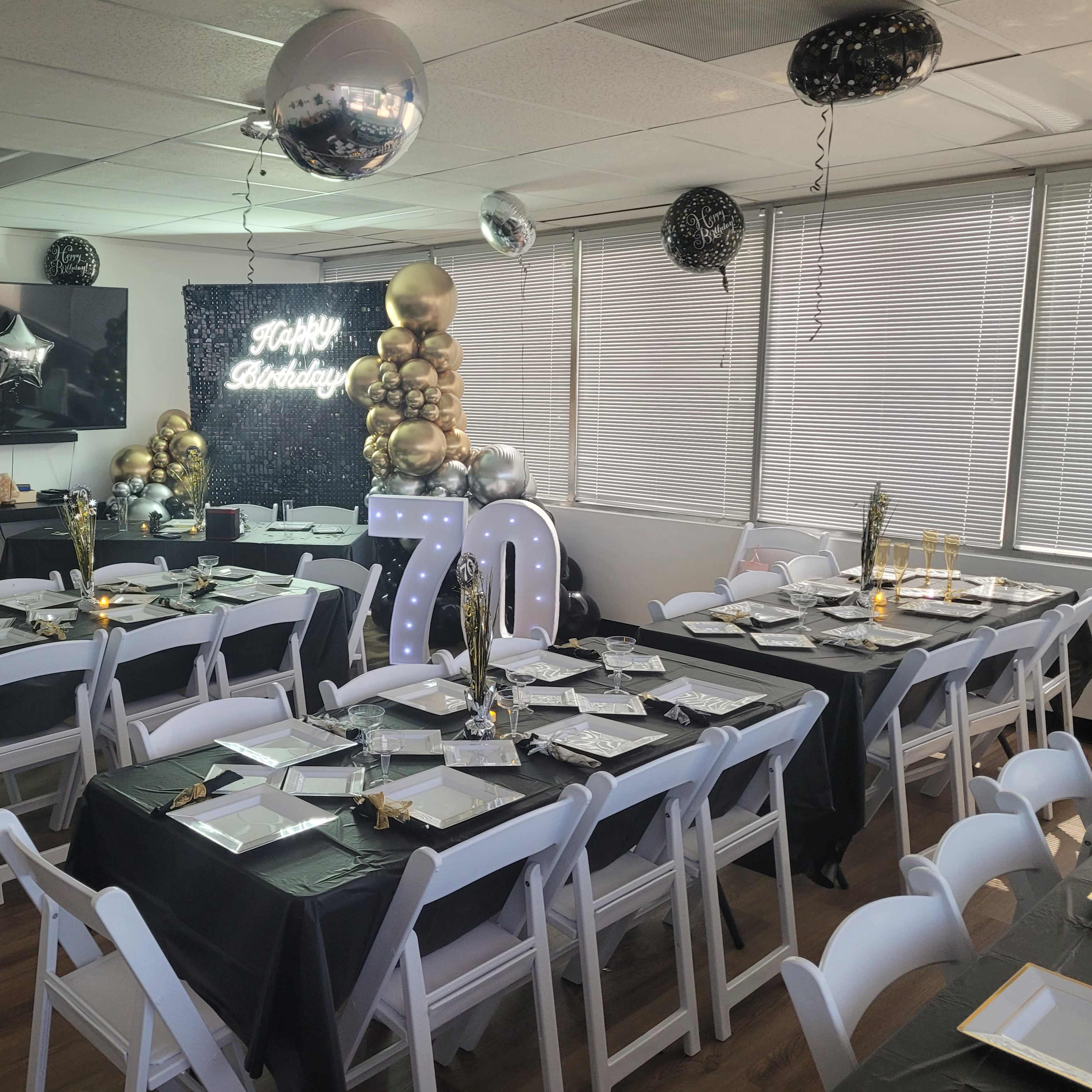 A decorated indoor party space set up for a 70th birthday celebration, featuring black and gold table settings, balloons, and a banner.