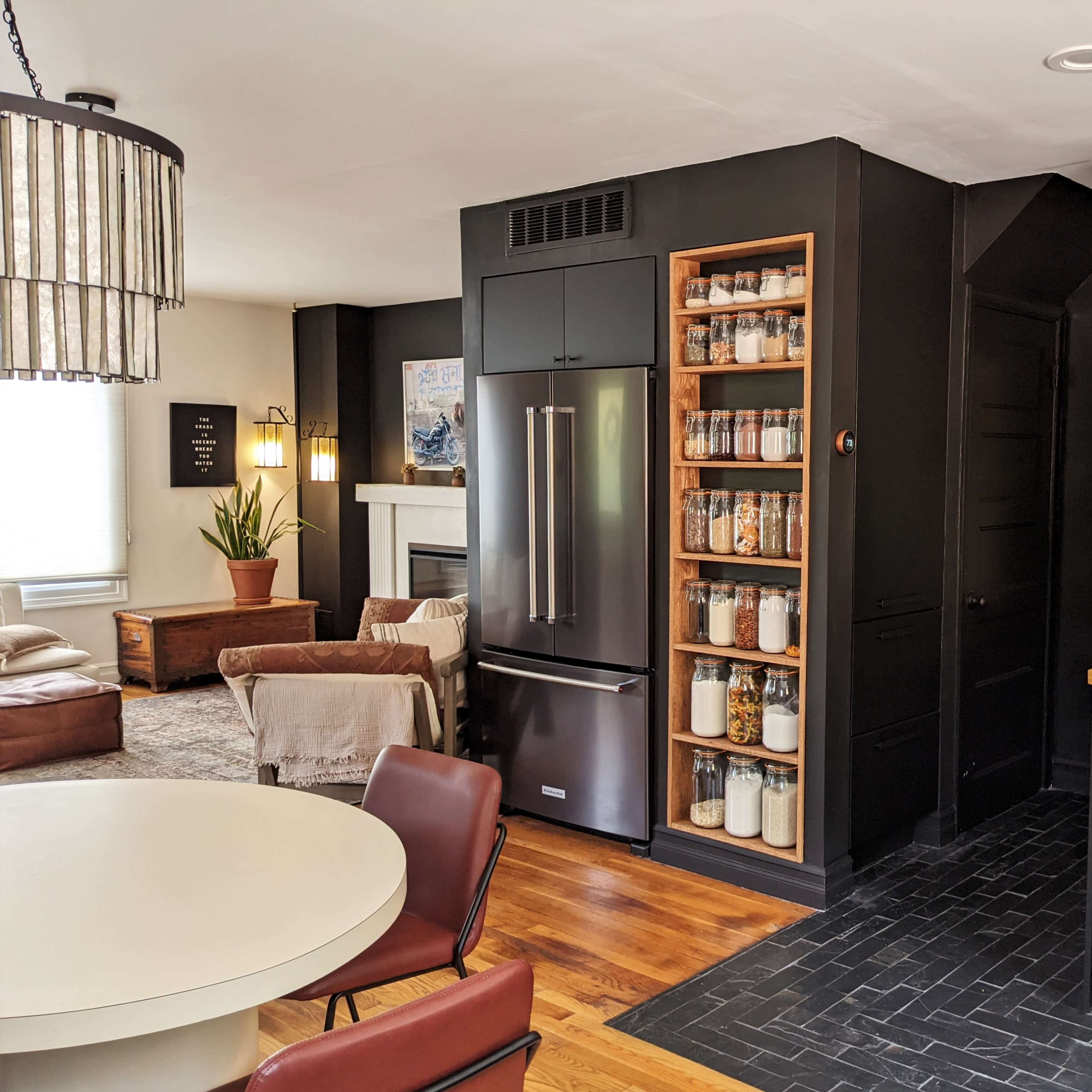 A modern kitchen and dining area with a dark cabinetry design, a refrigerator, and open shelving displaying jars and plants.