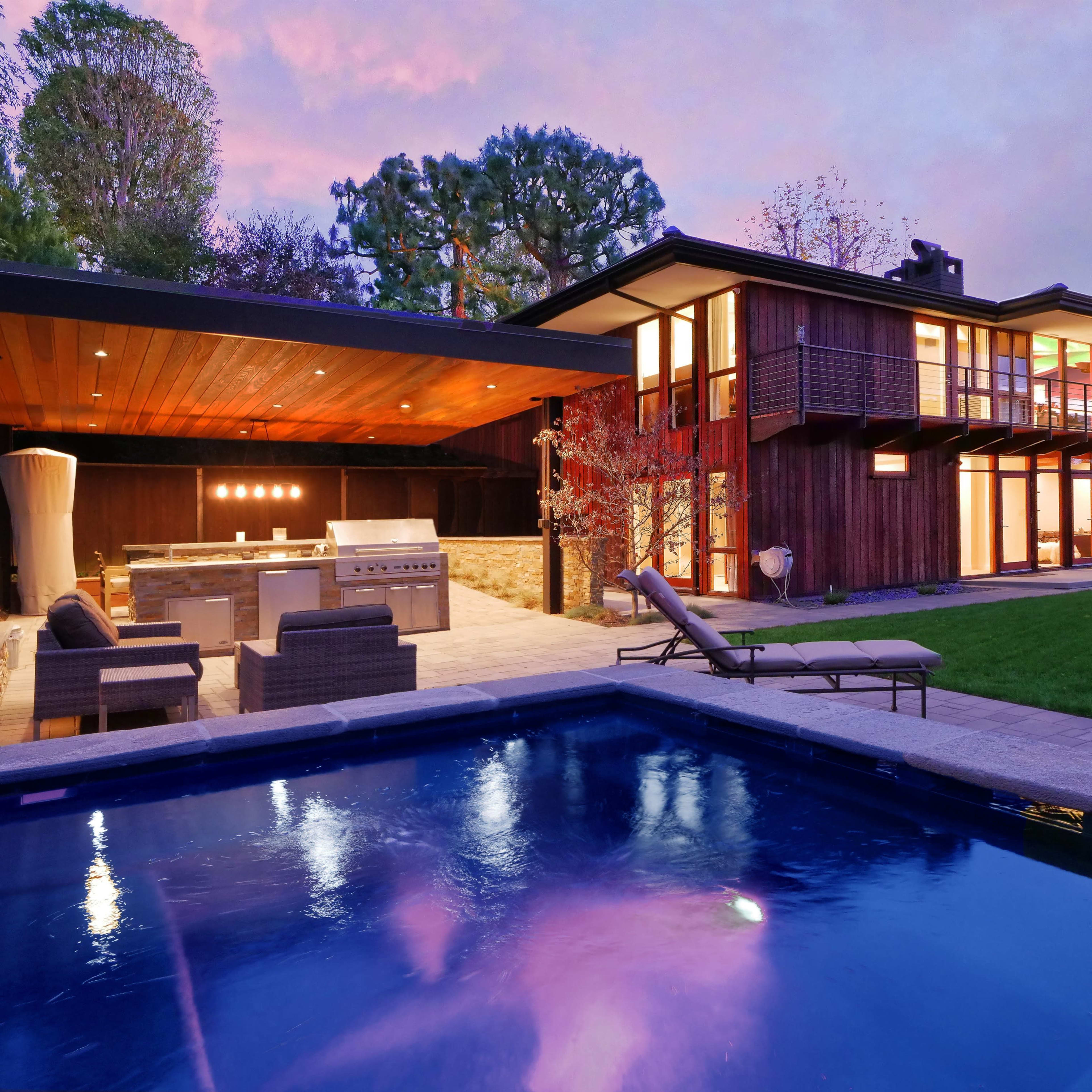 A modern house with large glass windows and a patio featuring an outdoor kitchen is set beside a swimming pool at dusk.