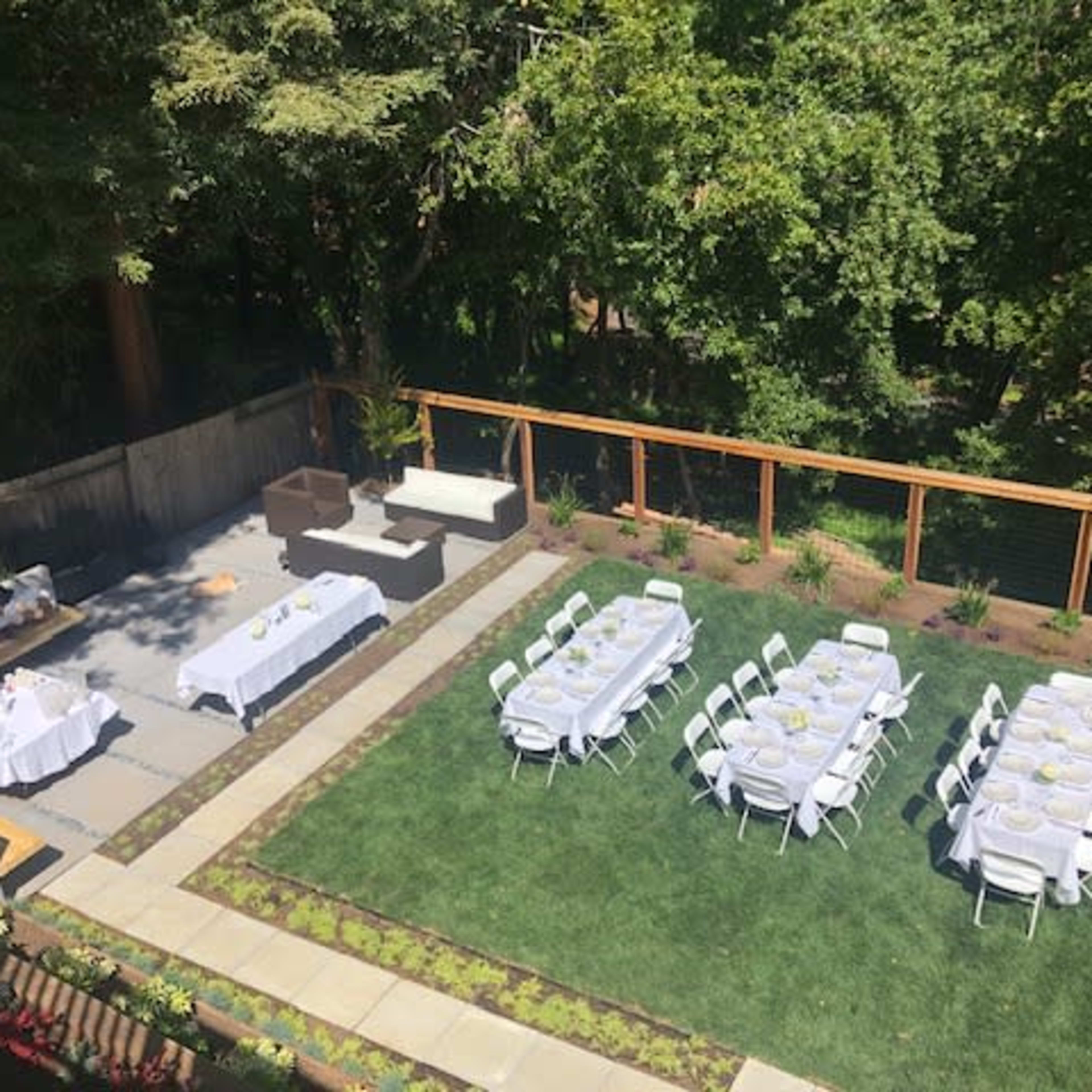 An outdoor event space with neatly arranged tables and chairs set on a lawn, surrounded by greenery and a wooden fence.
