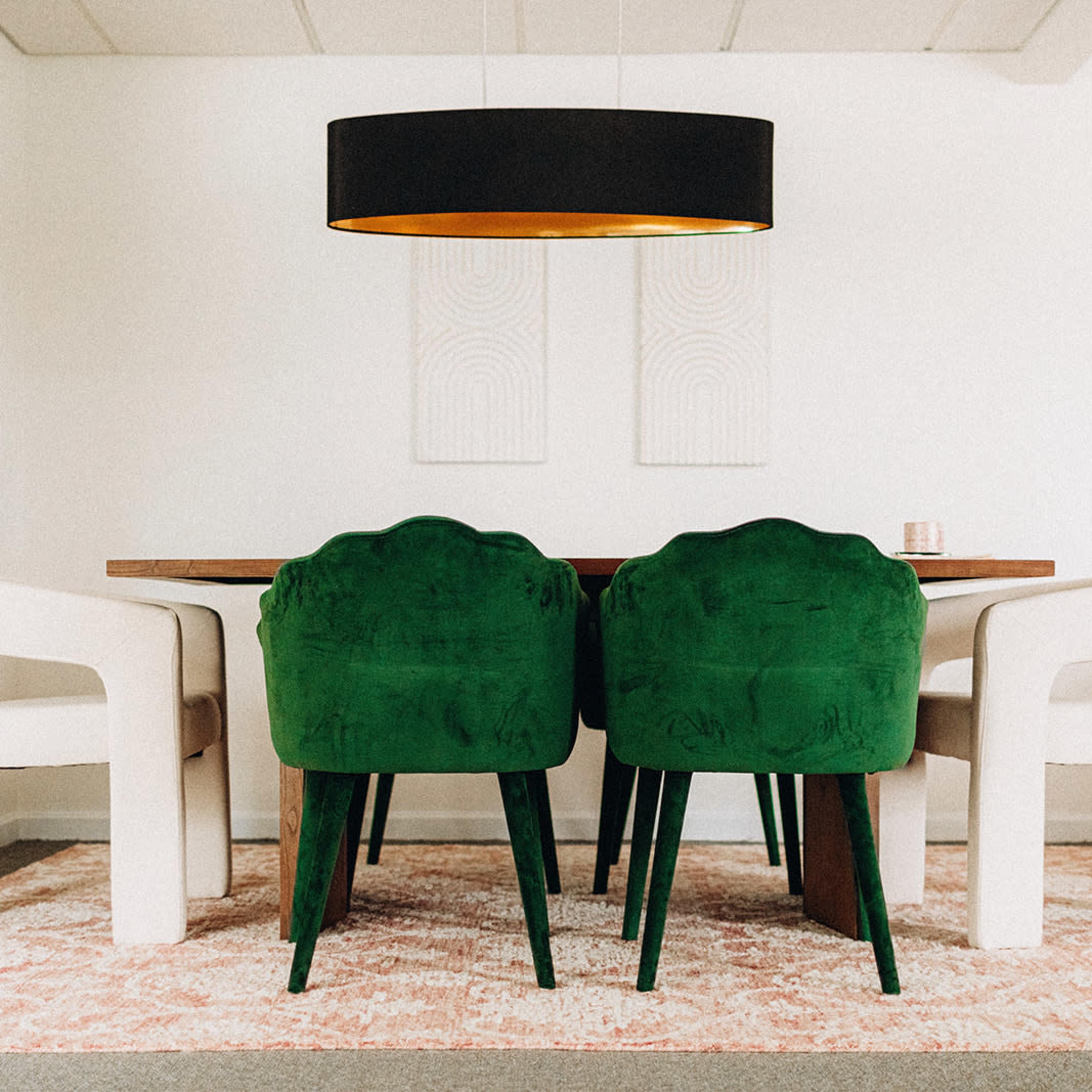 A dining area features a wooden table surrounded by two green chairs and two cream-colored armchairs, with a large pendant light above and a light pink rug underneath.
