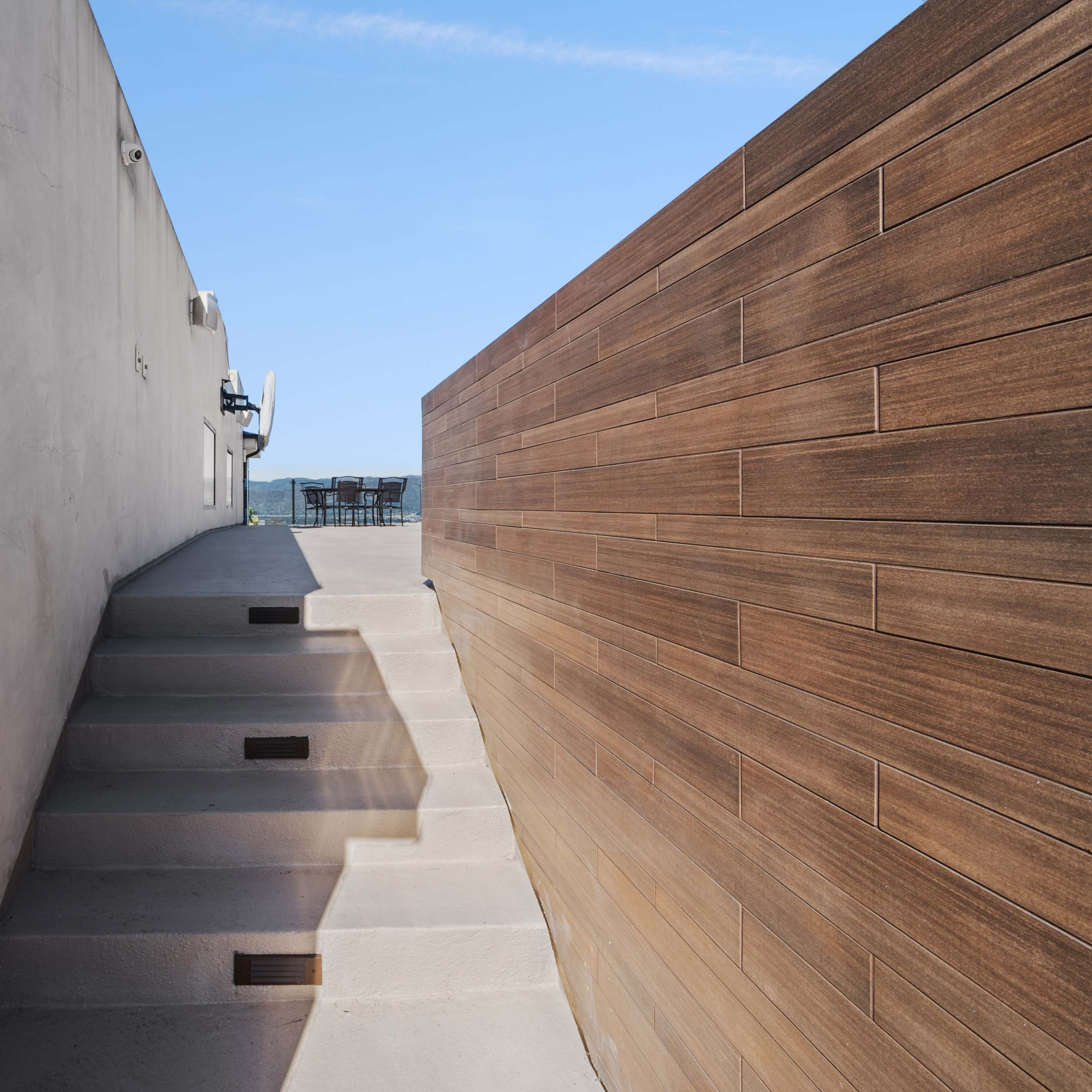 A narrow stairway leads up between a textured concrete wall and a wooden paneling toward a clear blue sky.
