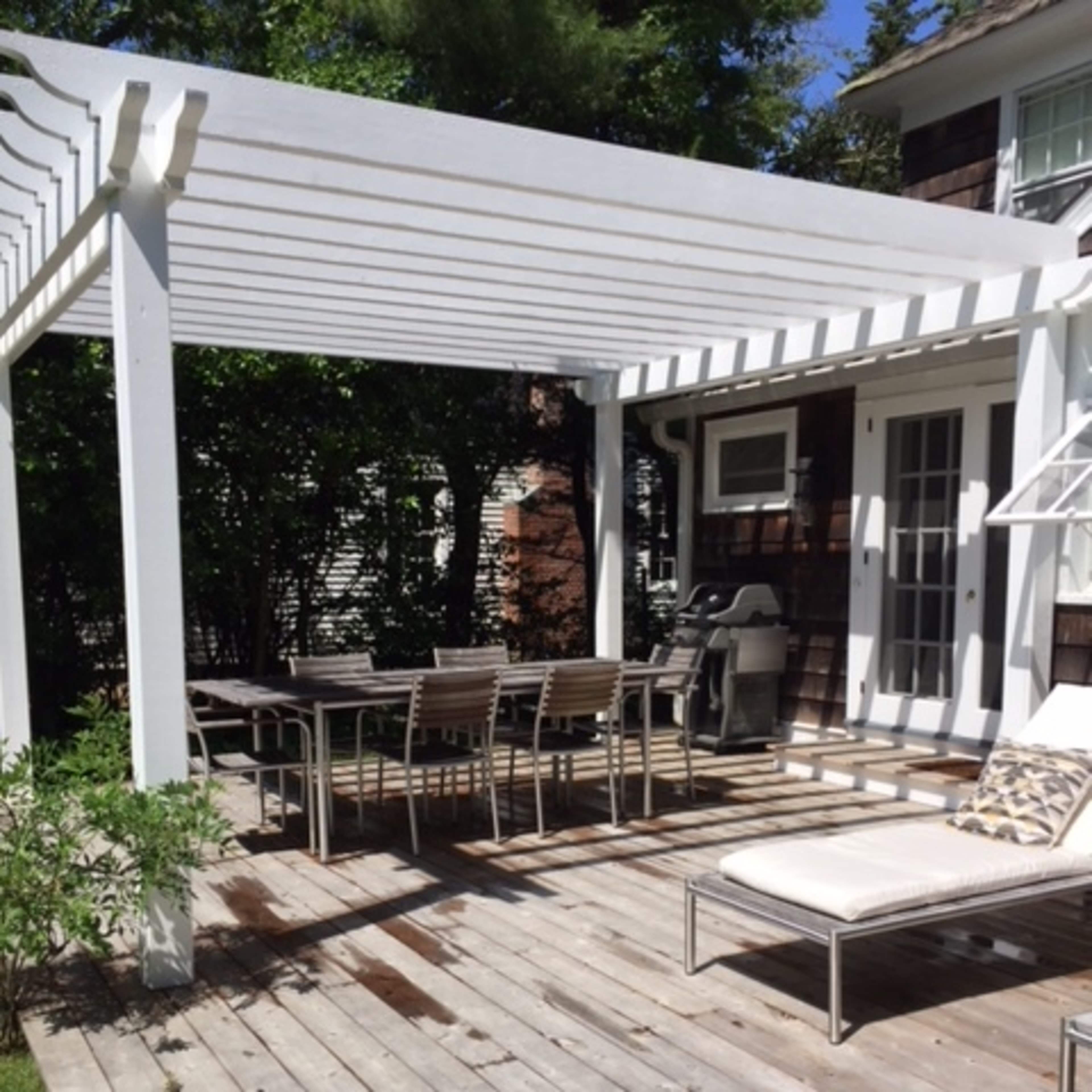 A wooden deck with a table and chairs situated under a white pergola, adjacent to a house.