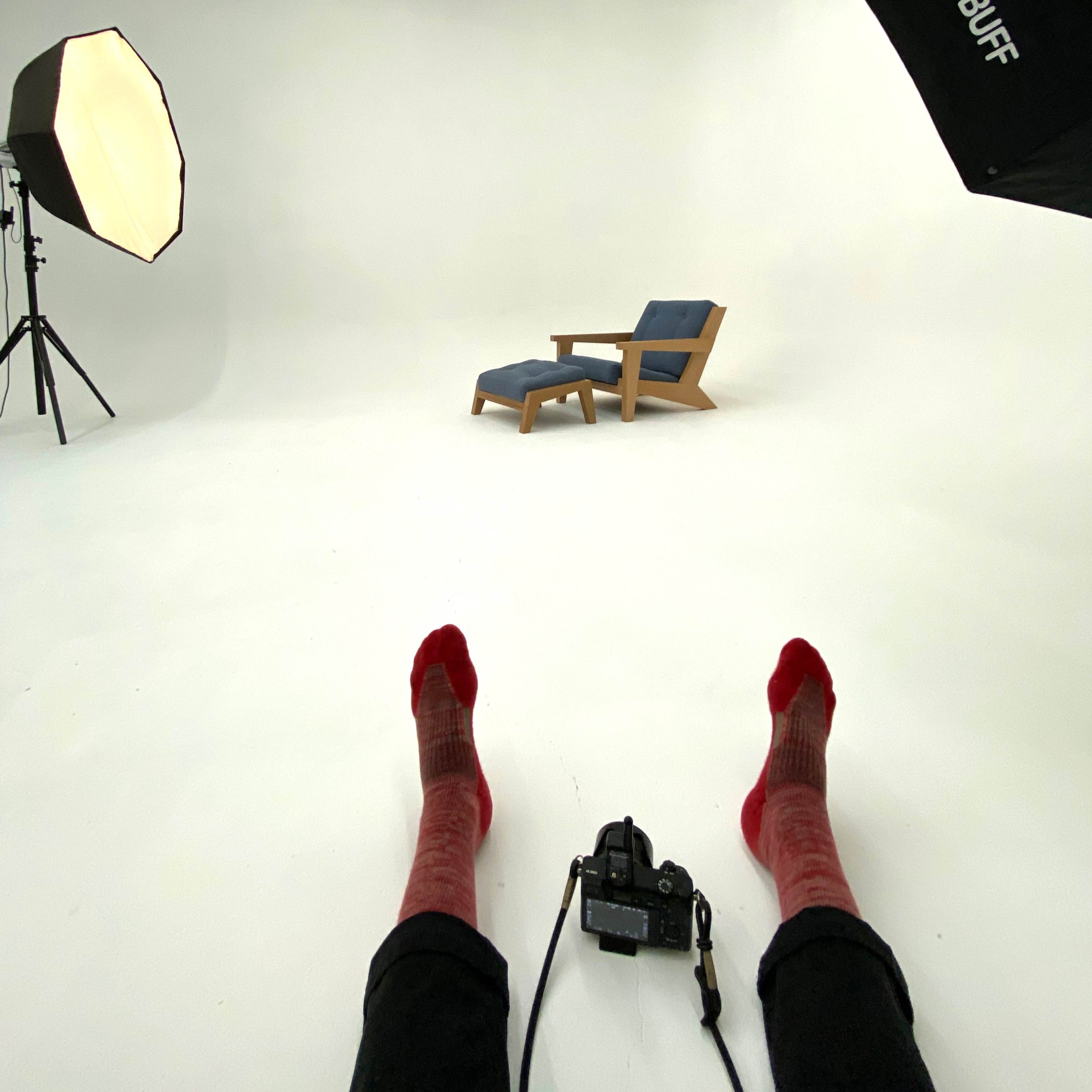 A person sits on the floor of a photography studio, wearing red socks, with a camera in front of a blue chair and lighting equipment.