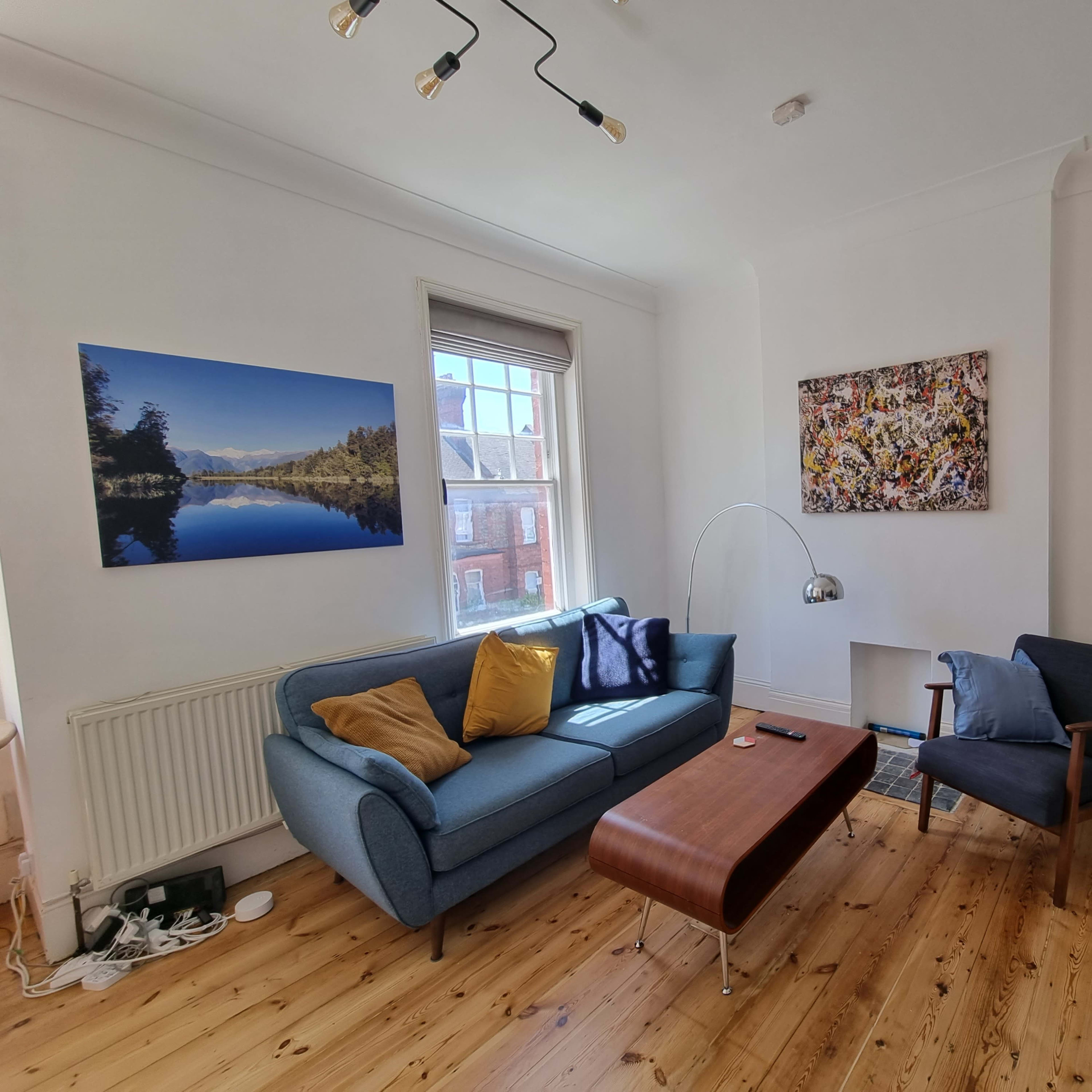 A bright living room with a blue sofa, a wooden coffee table, and a large window, featuring artwork on the walls and a chess set on a side table.