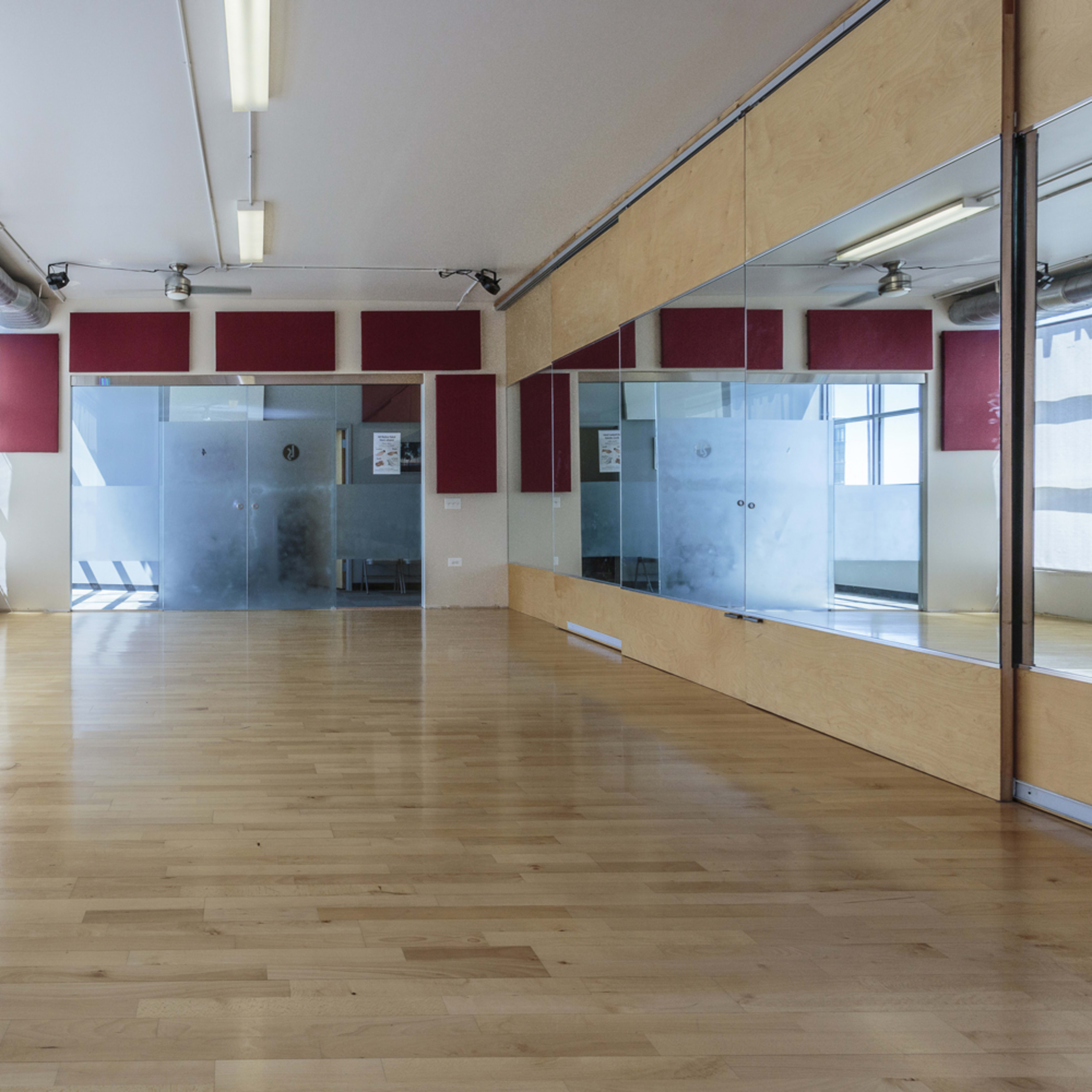 An empty dance studio with wooden flooring, large windows letting in natural light, and a mirrored wall.