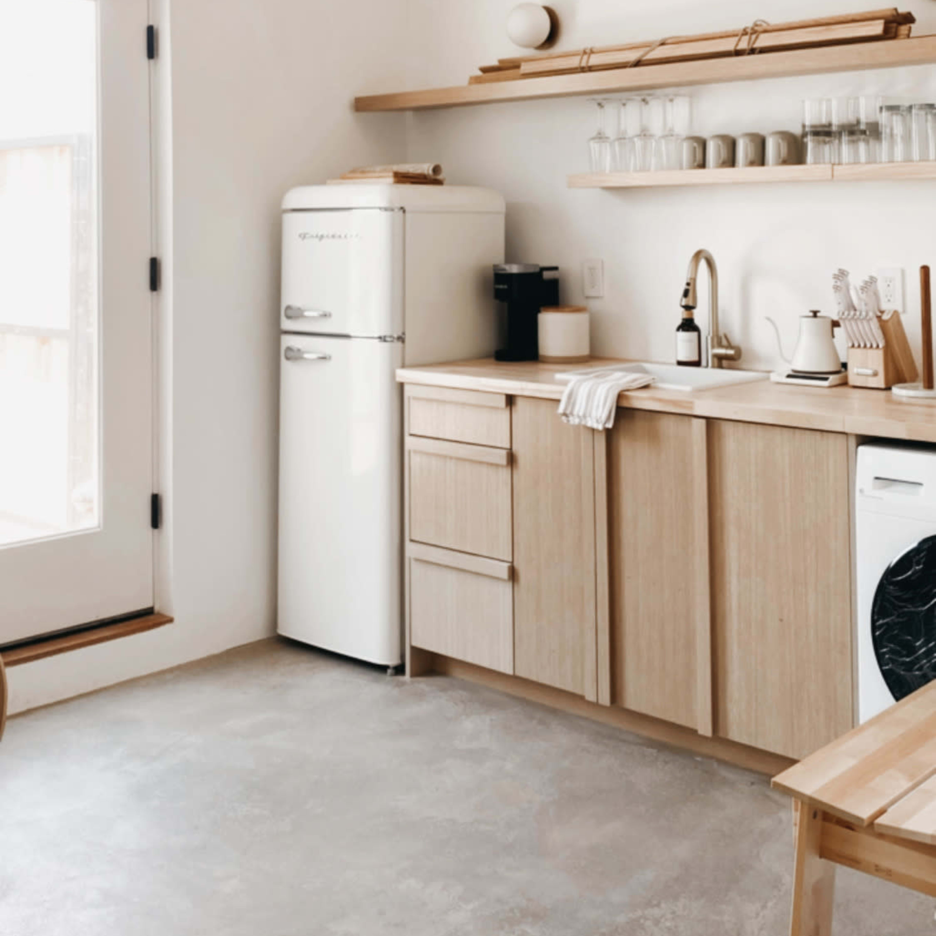 A modern kitchen features a white refrigerator, wooden cabinetry, and shelves with glassware and kitchen tools, illuminated by natural light from a nearby door.
